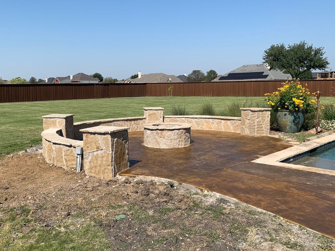 Stone fire pit and seating area in a backyard, beside a pool and lawn, with a wooden fence in the background.