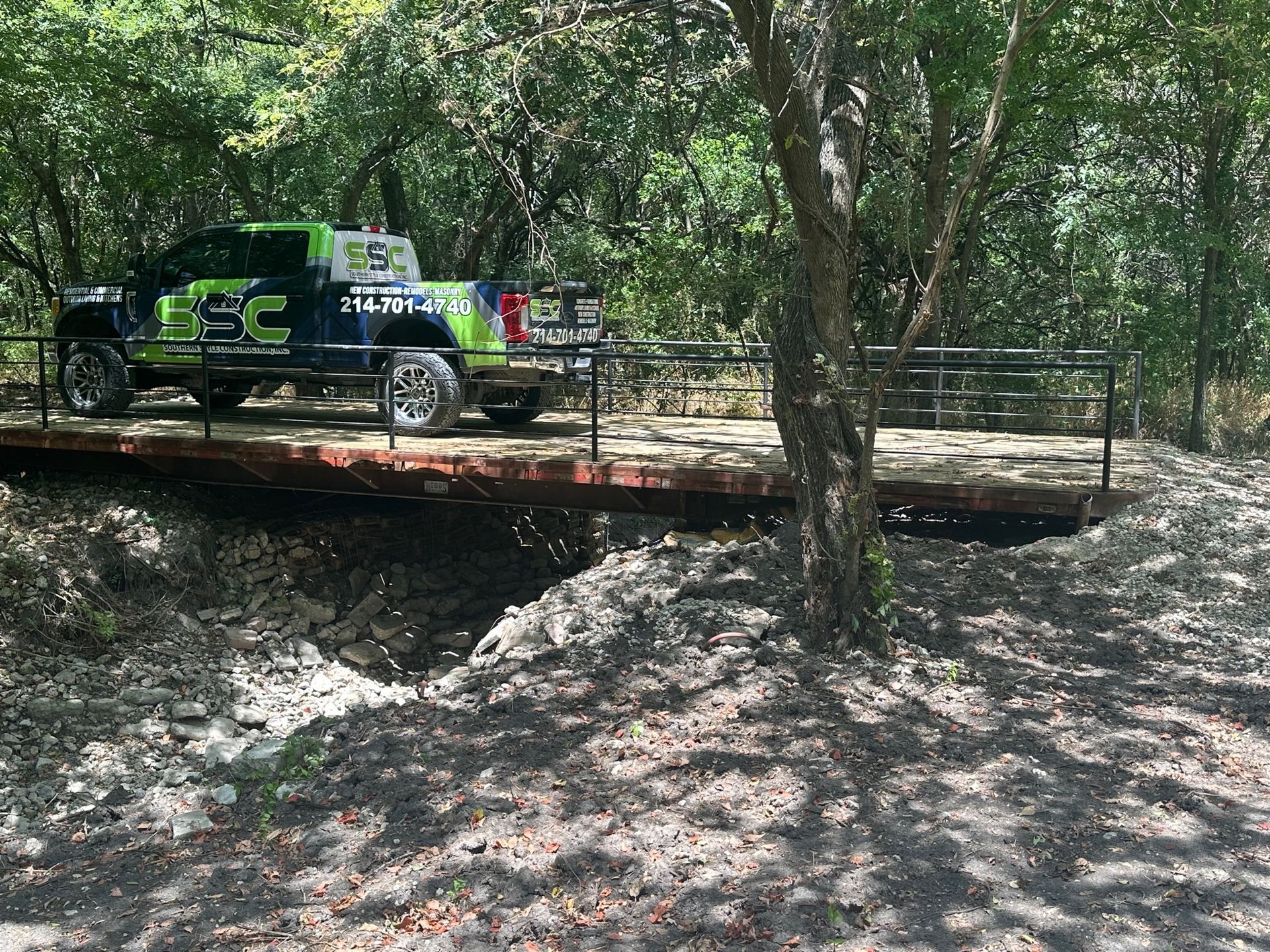 Green and black pickup truck on a makeshift bridge over a dry creek bed in a wooded area.