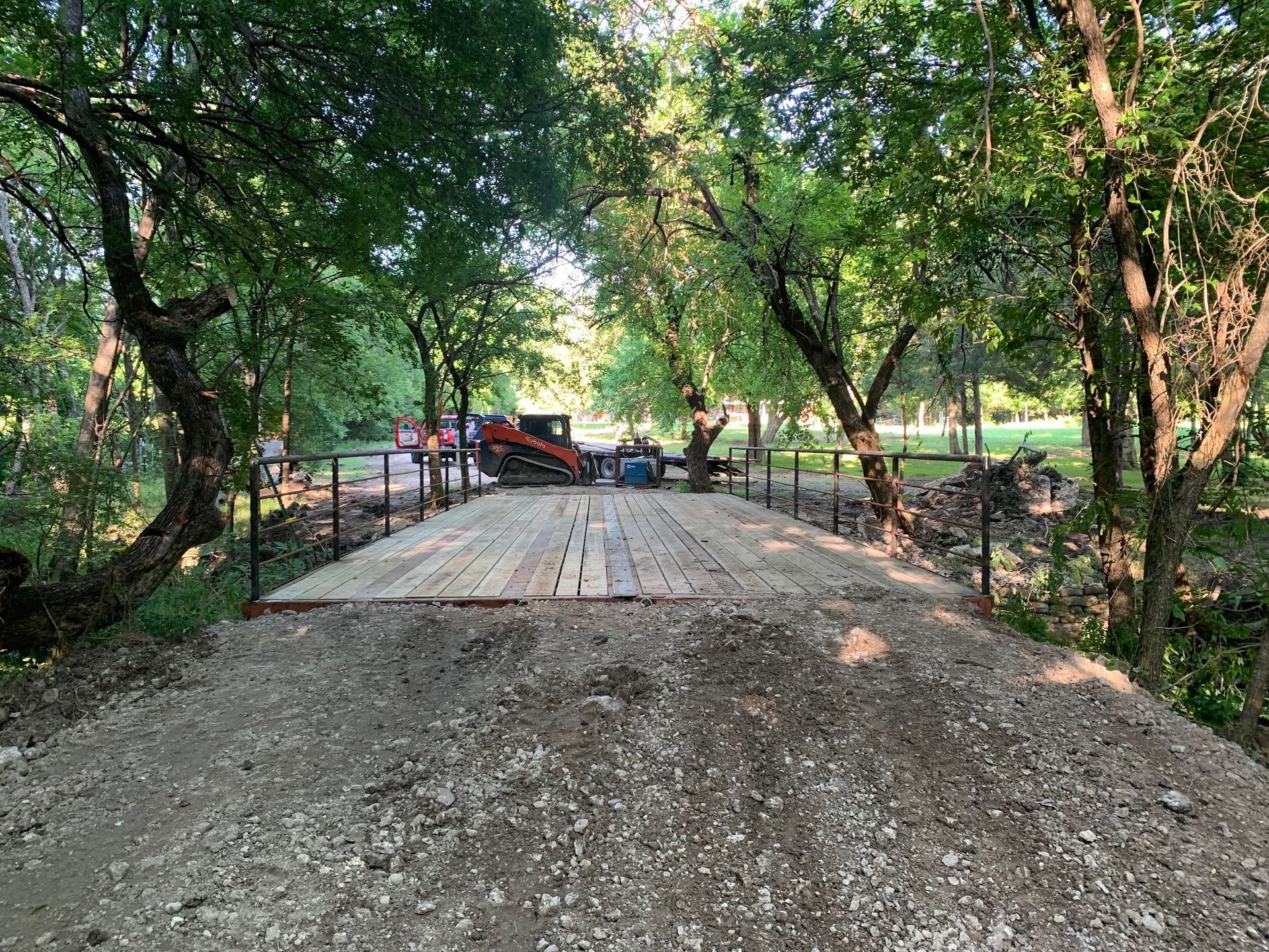 A new gravel bridge with wood planks, constructed in a wooded area. Heavy equipment visible in the distance.