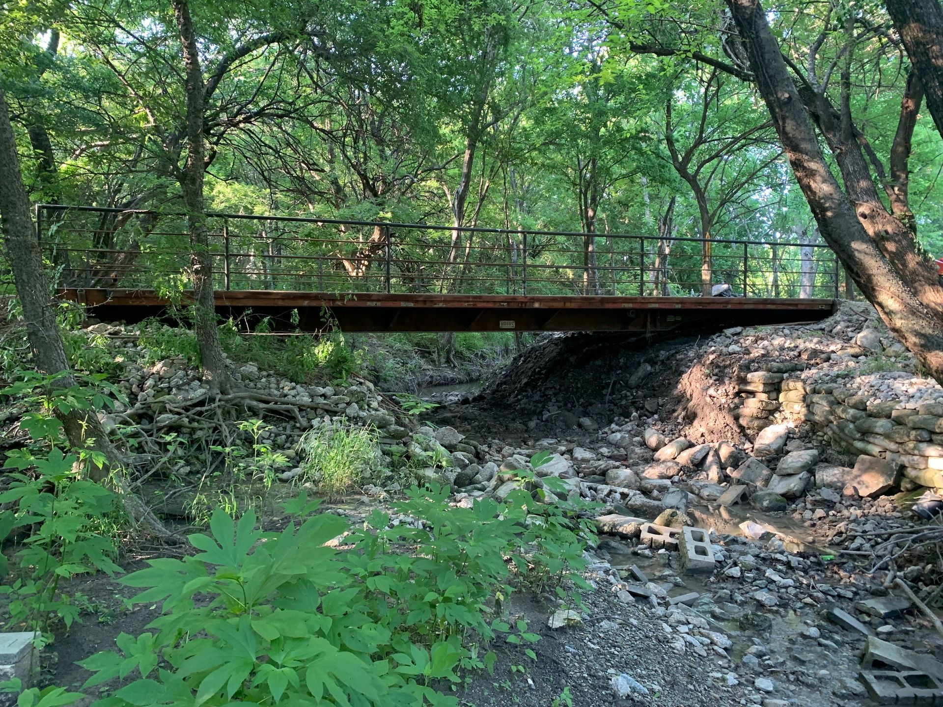 Metal bridge over a dry creek bed in a wooded area; person stands on bridge.