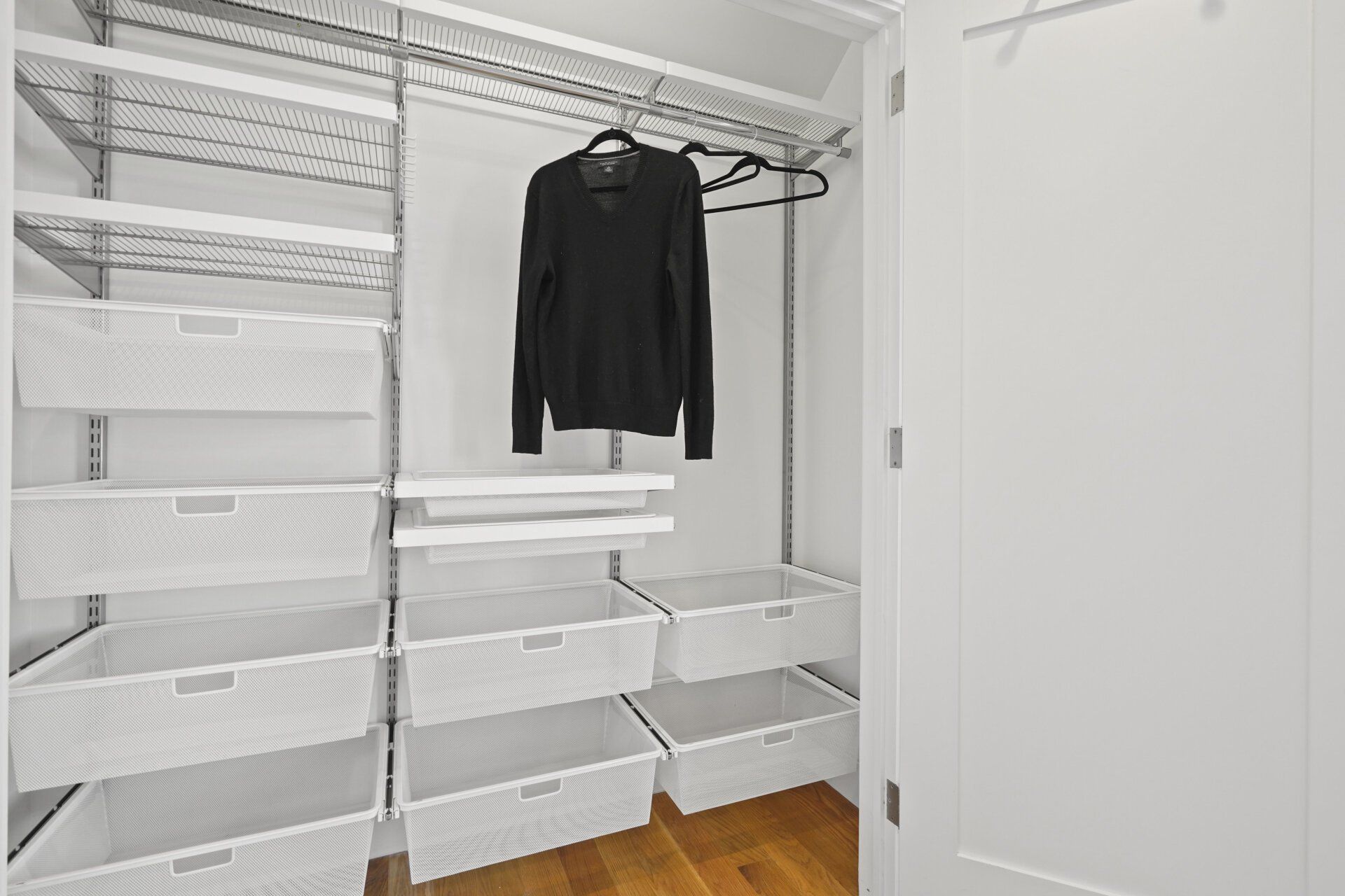A white closet with open wire drawers, a hanging sweater, and a door.