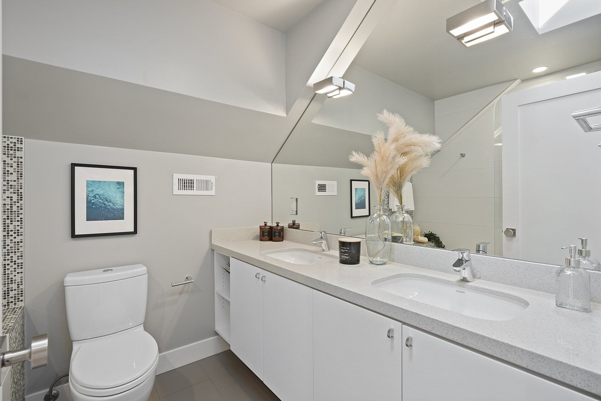 A bathroom with a white vanity, toilet, and a large mirror. Features include neutral walls, artwork, and a vase with pampas grass.