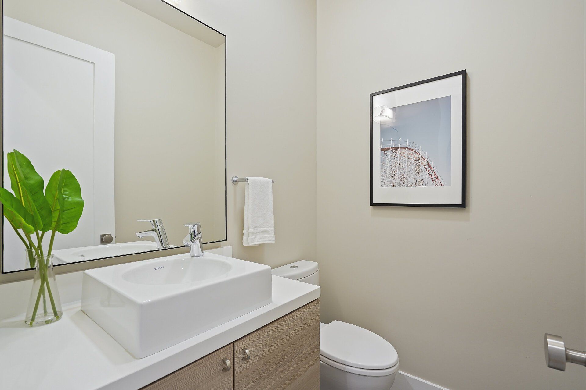 A bathroom with sink, large mirror, and framed art. Neutral walls, light wood vanity, and a green plant add visual interest.