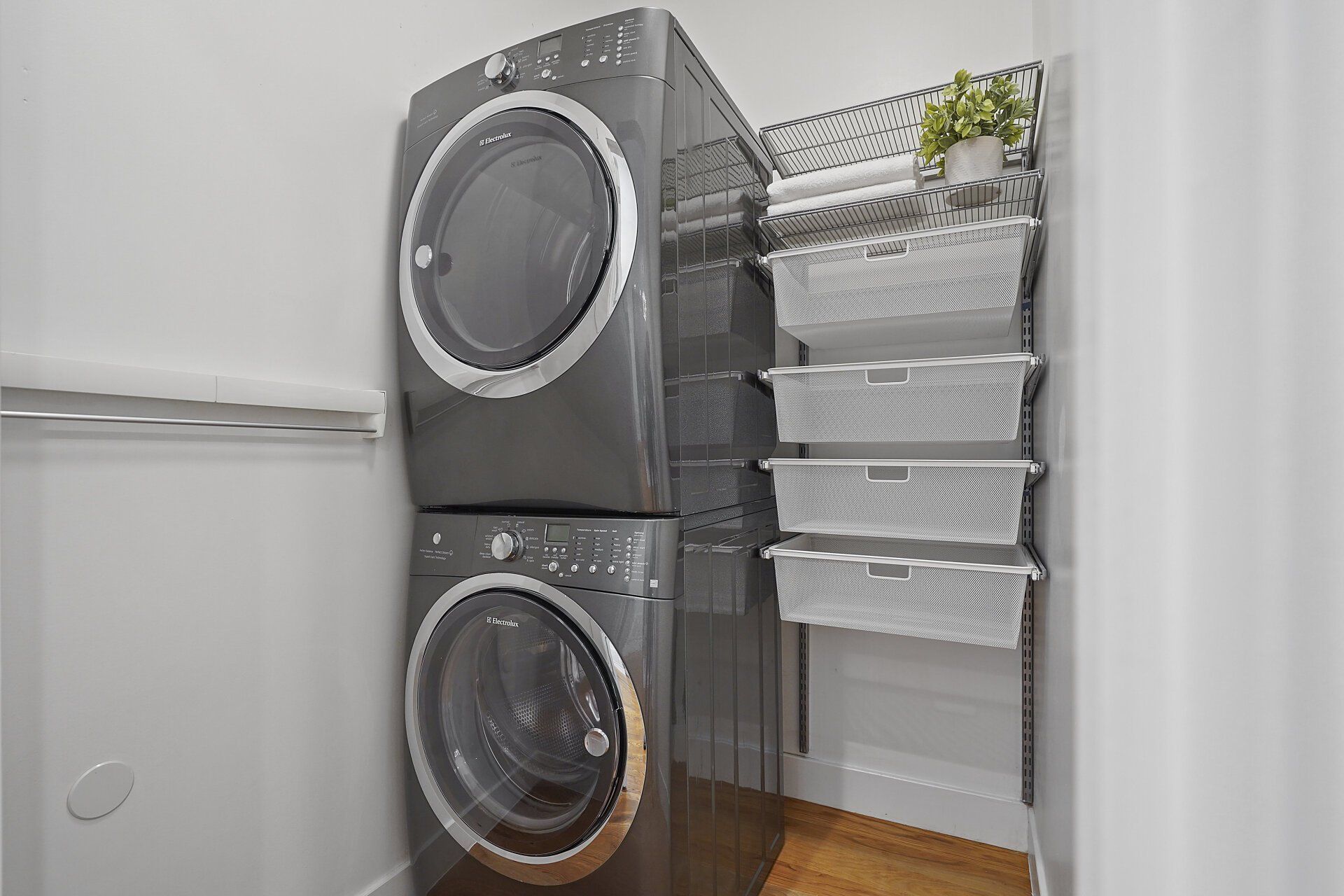 Stacked gray washer and dryer next to a wall-mounted shelving unit with four white bins for storage in a laundry room.