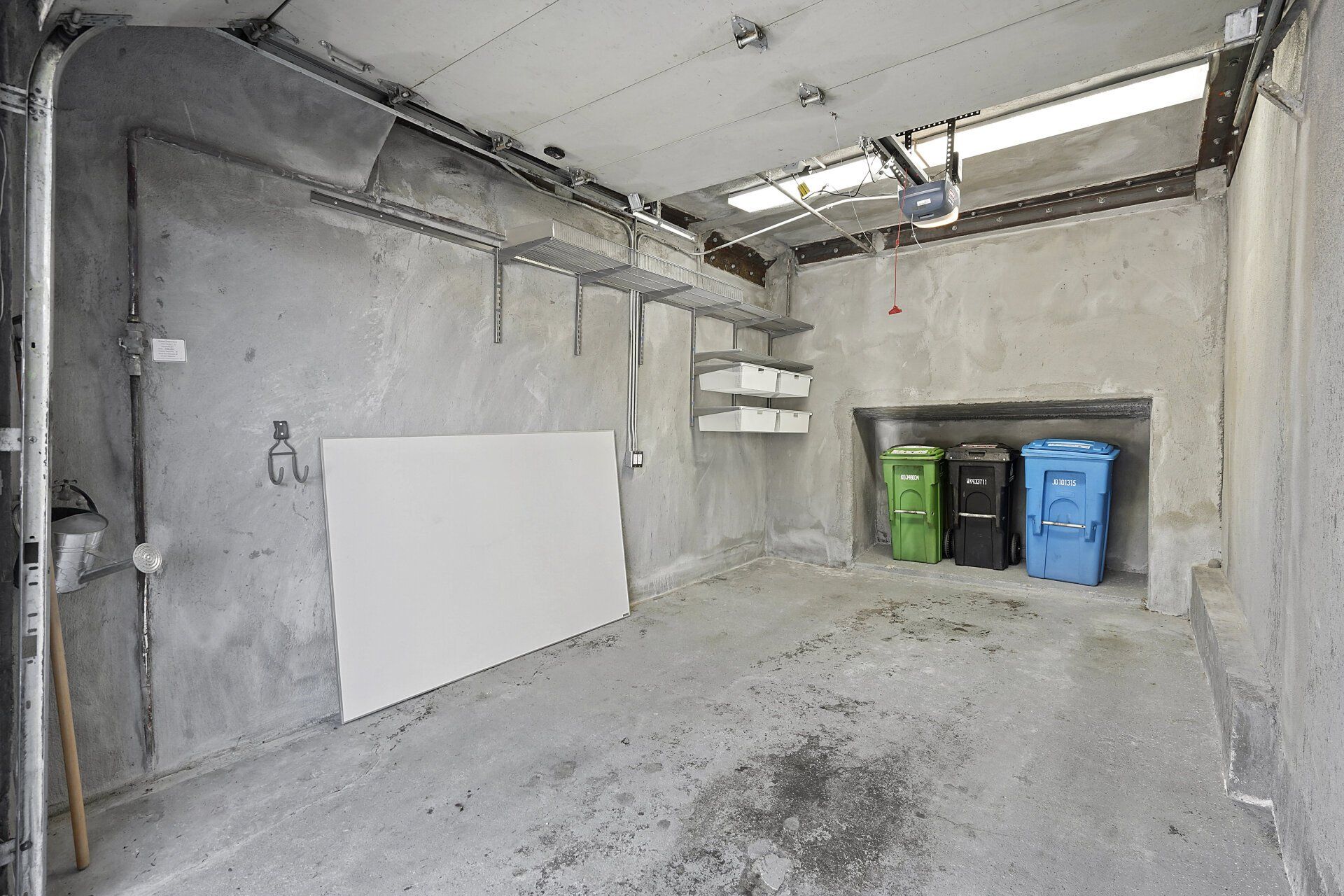 An empty concrete garage with shelves, a large white board, and three trash bins.