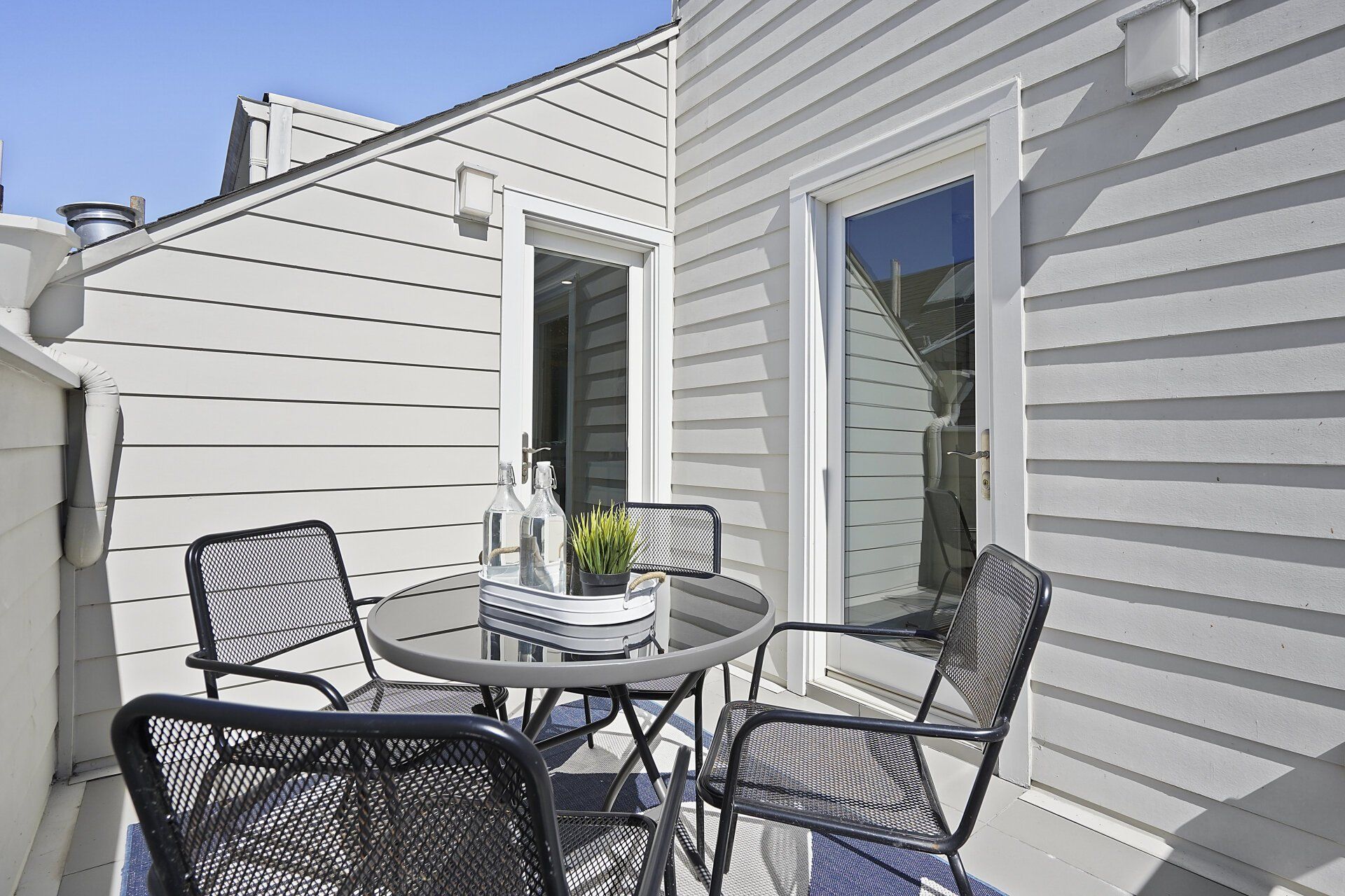 Outdoor patio with gray siding and a glass-topped table with four chairs. Two doors lead inside.