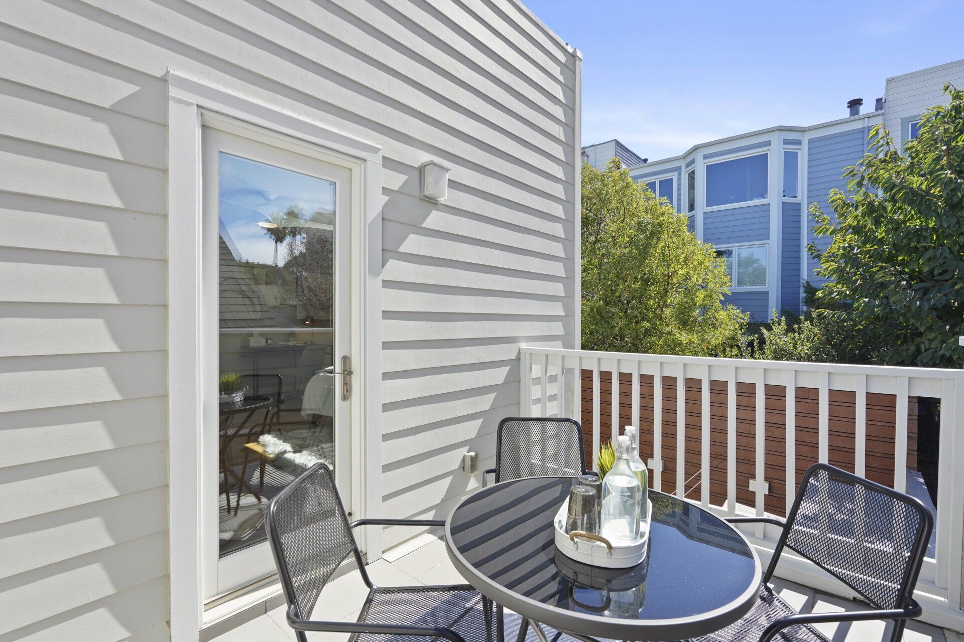 A balcony with a glass-topped table, chairs, and a glass door. The balcony is bordered by a white railing and grey siding.