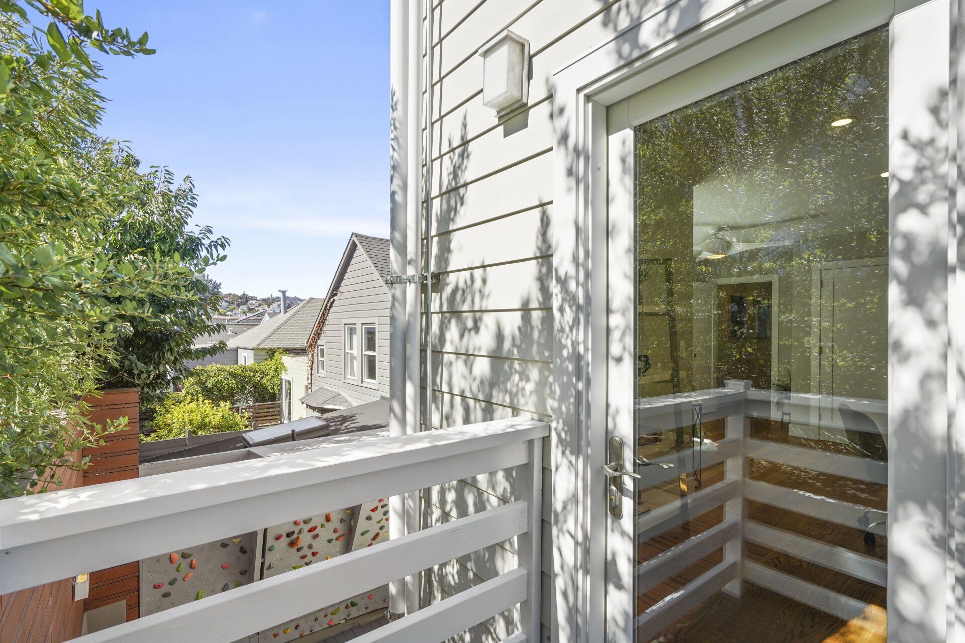 Balcony with white railing, glass door, and view of a neighborhood with houses and trees. Sunlight casts shadows.