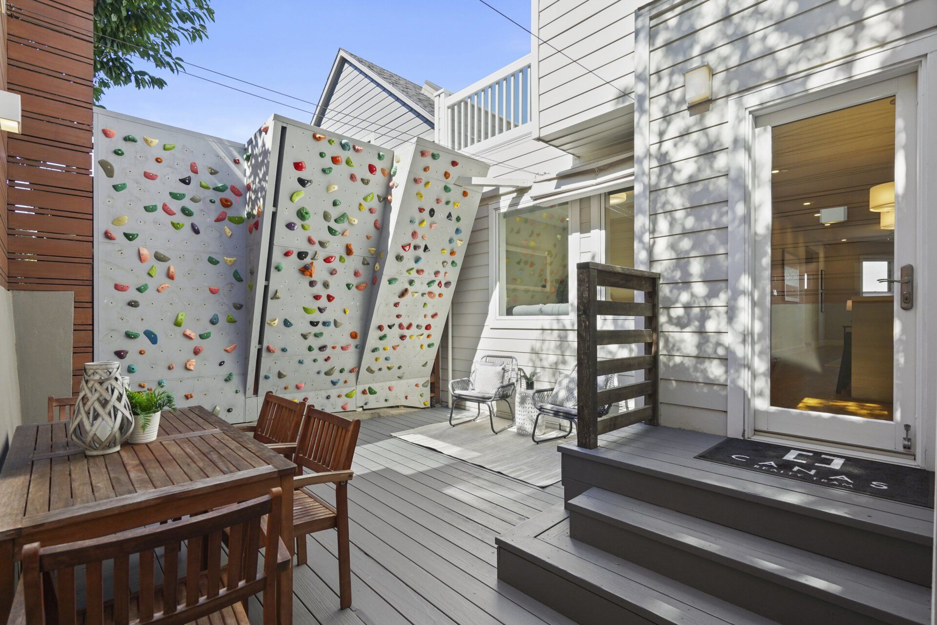 A backyard patio with a climbing wall, a wooden table, and a door to a house. The climbing wall has various colored holds.