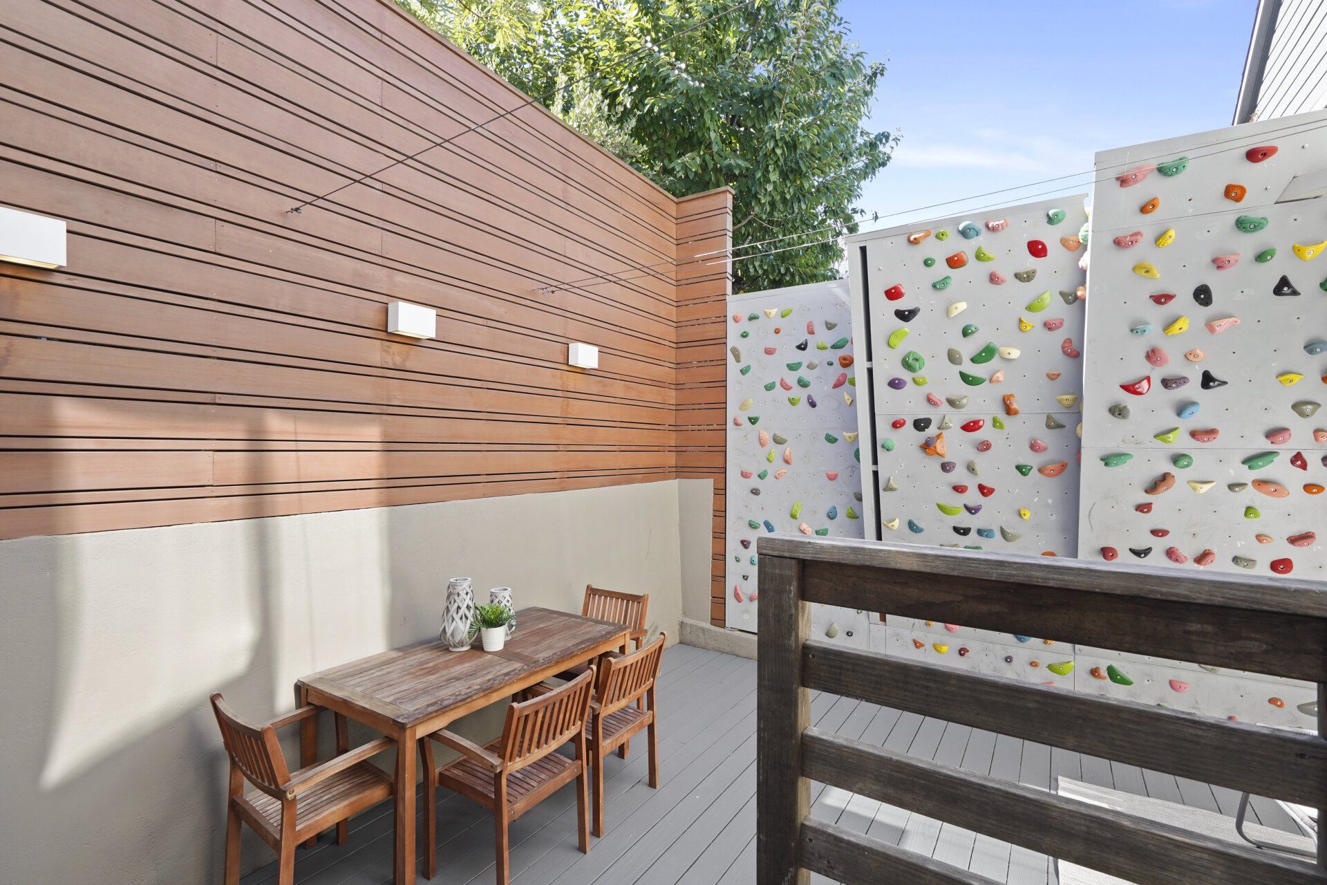 Outdoor patio with wooden table and chairs, bordered by walls: one horizontal wood paneling, the other with colorful climbing holds.