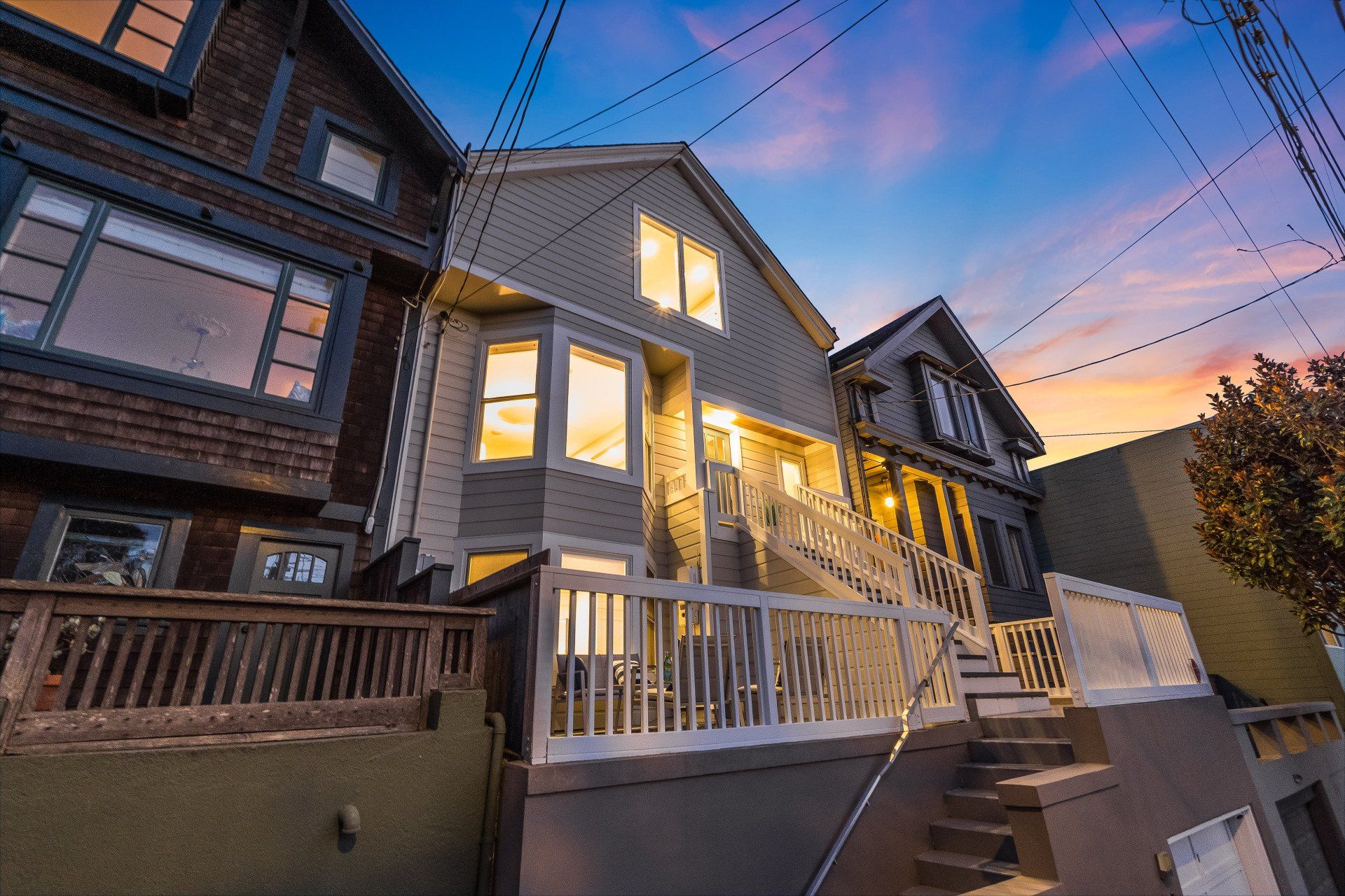 Row of houses at dusk. The center house, painted grey with a white railing, has lights on inside. Steps lead up to its entrance.