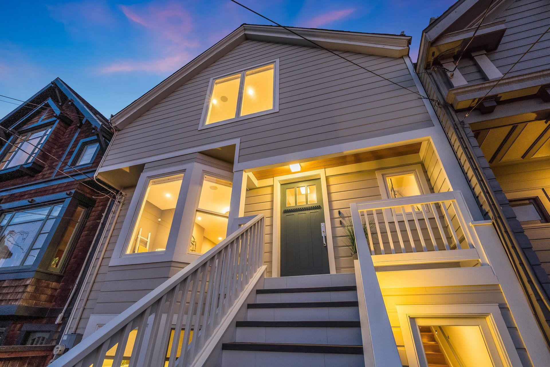 Beige two-story house with a dark colored door and white trim. The house has stairs leading up to the entrance with warm interior lighting.