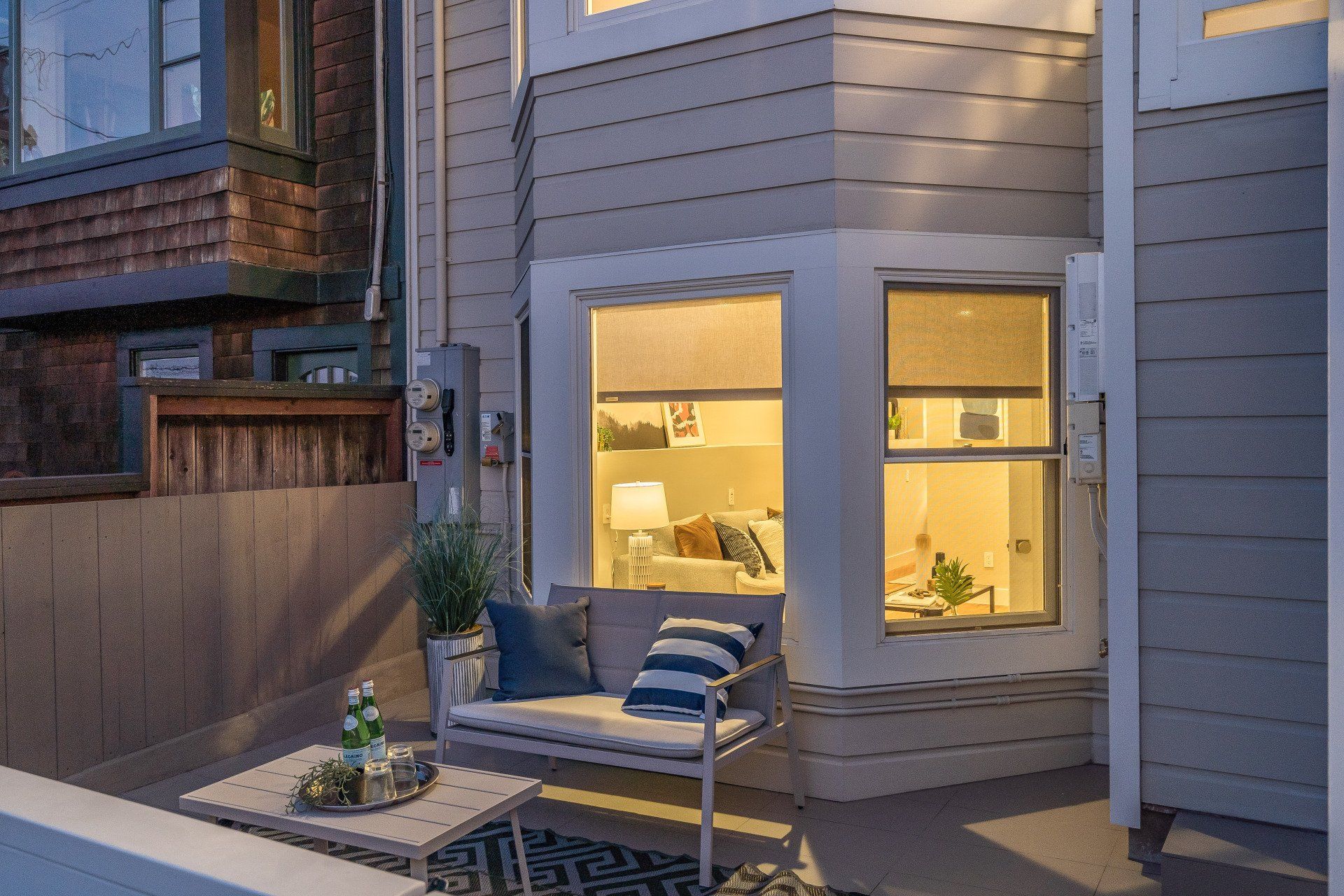 Outdoor seating area with a gray couch and small table, illuminated by interior lights shining through the windows of a building.