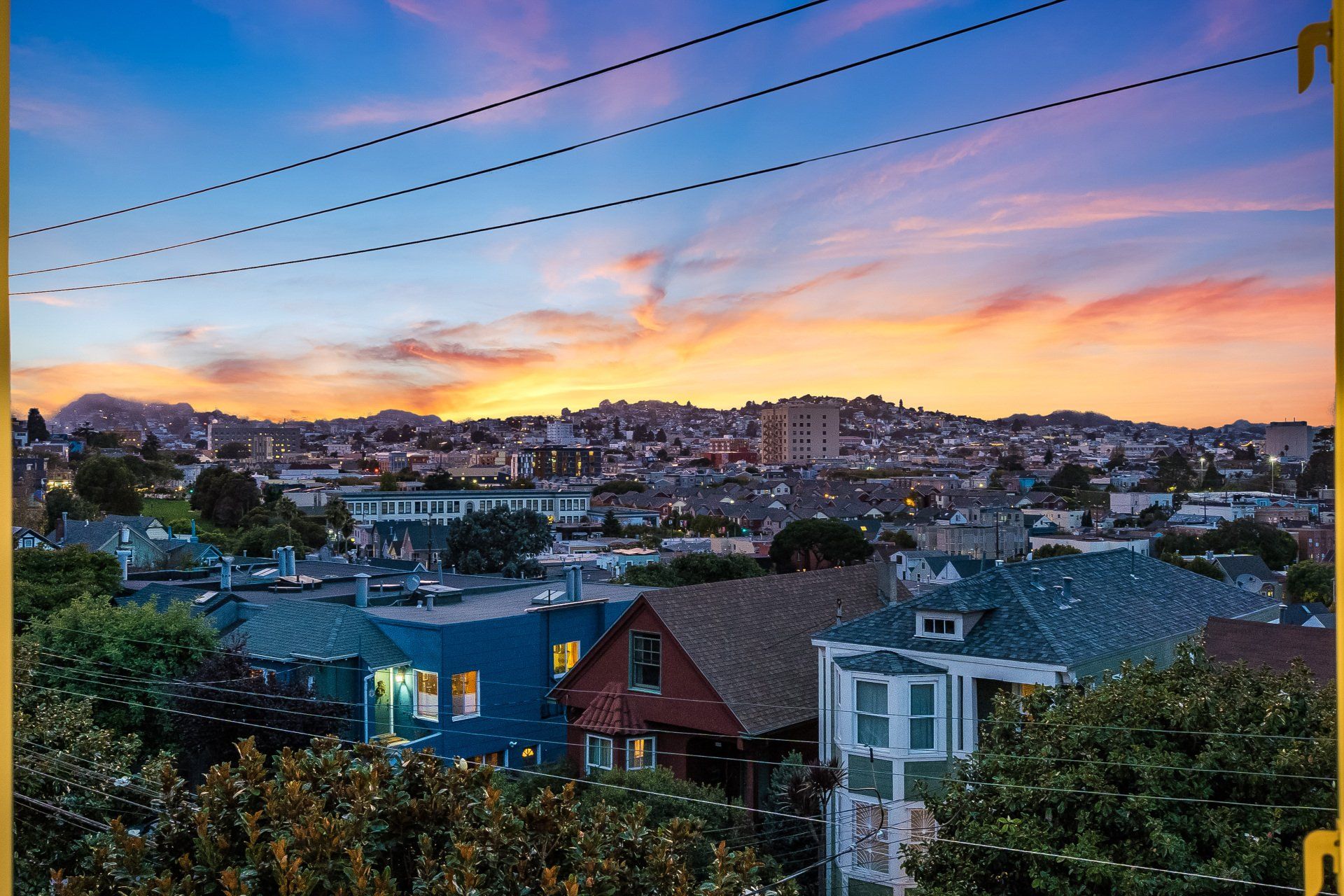 Cityscape at sunset: Houses and trees in the foreground, with a colorful sky and distant buildings silhouetted on hills.