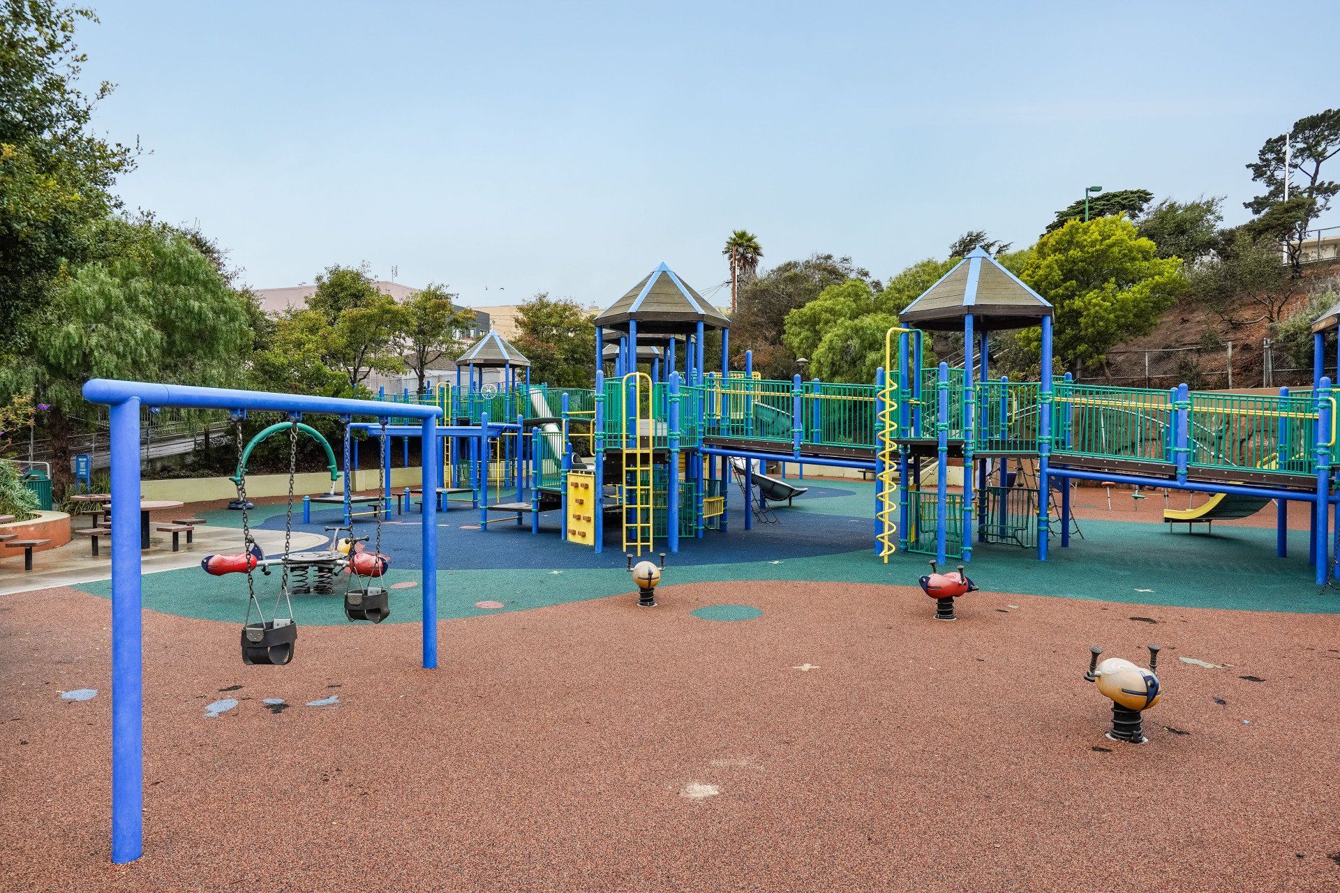 Playground with blue and green equipment, including swings, slides, and climbing structures. Brown ground and trees surround the play area.
