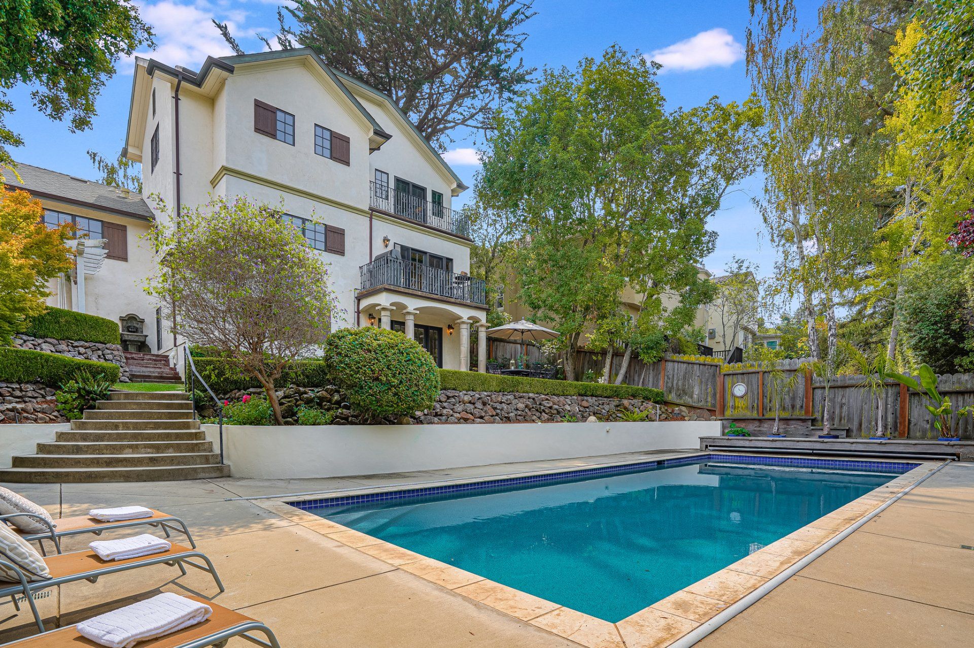 A large, light-colored house with a balcony overlooks a pool and patio; trees and landscaping surround the property.
