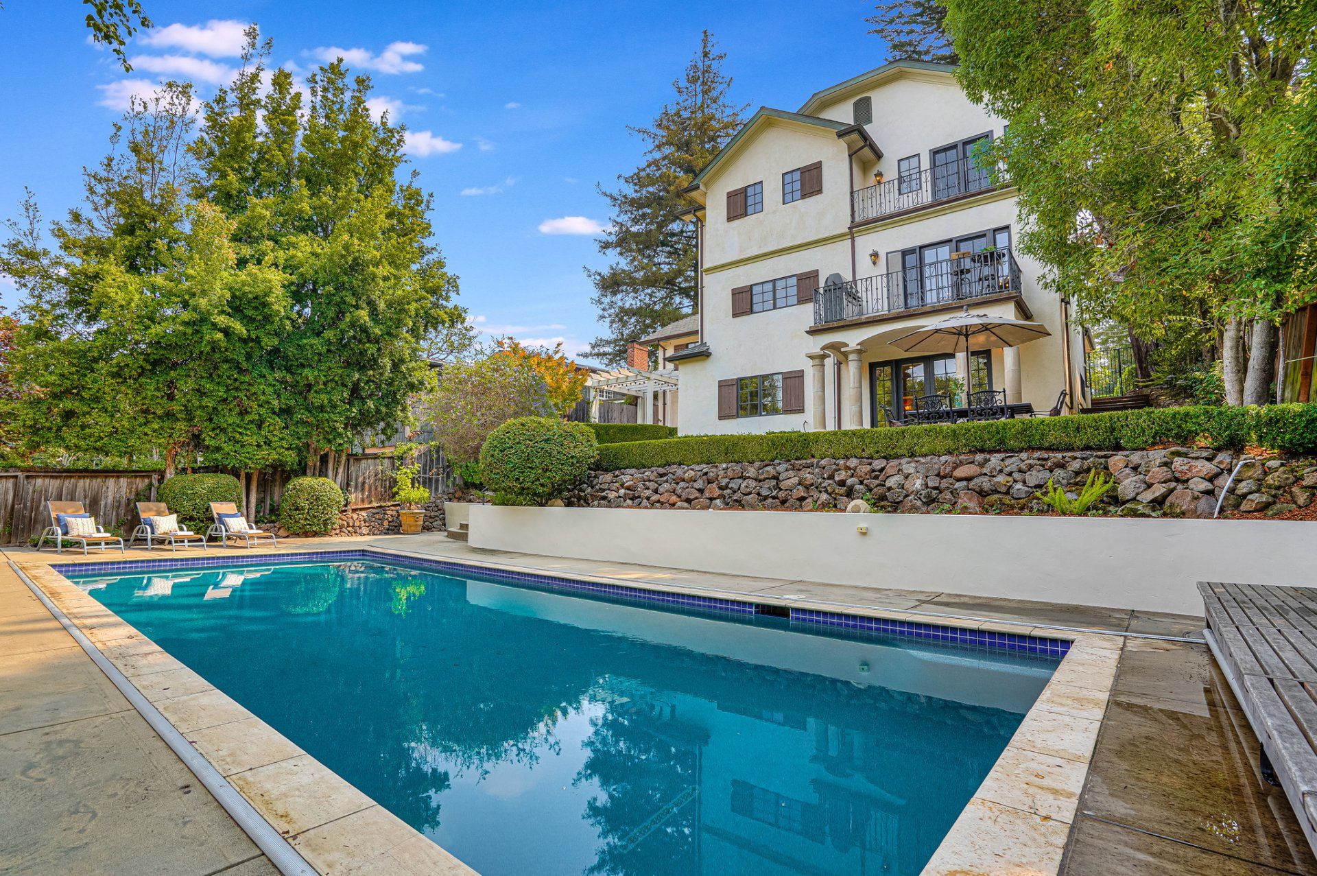 A large house with a swimming pool in its backyard under a blue sky. Lush greenery surrounds the home.