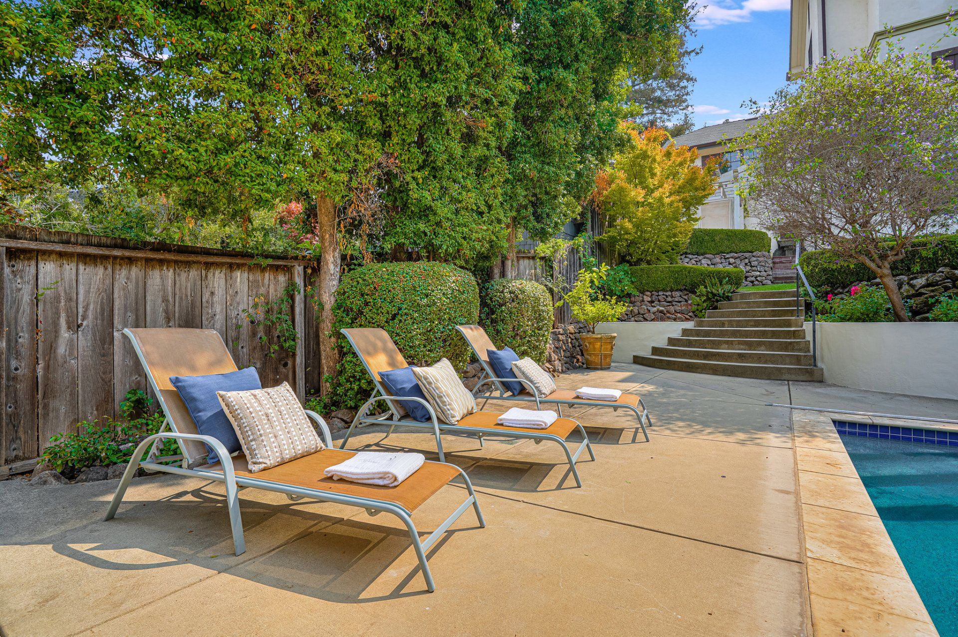 Lounge chairs by a pool, in a backyard setting. The chairs are tan with blue and cream pillows, and white towels.