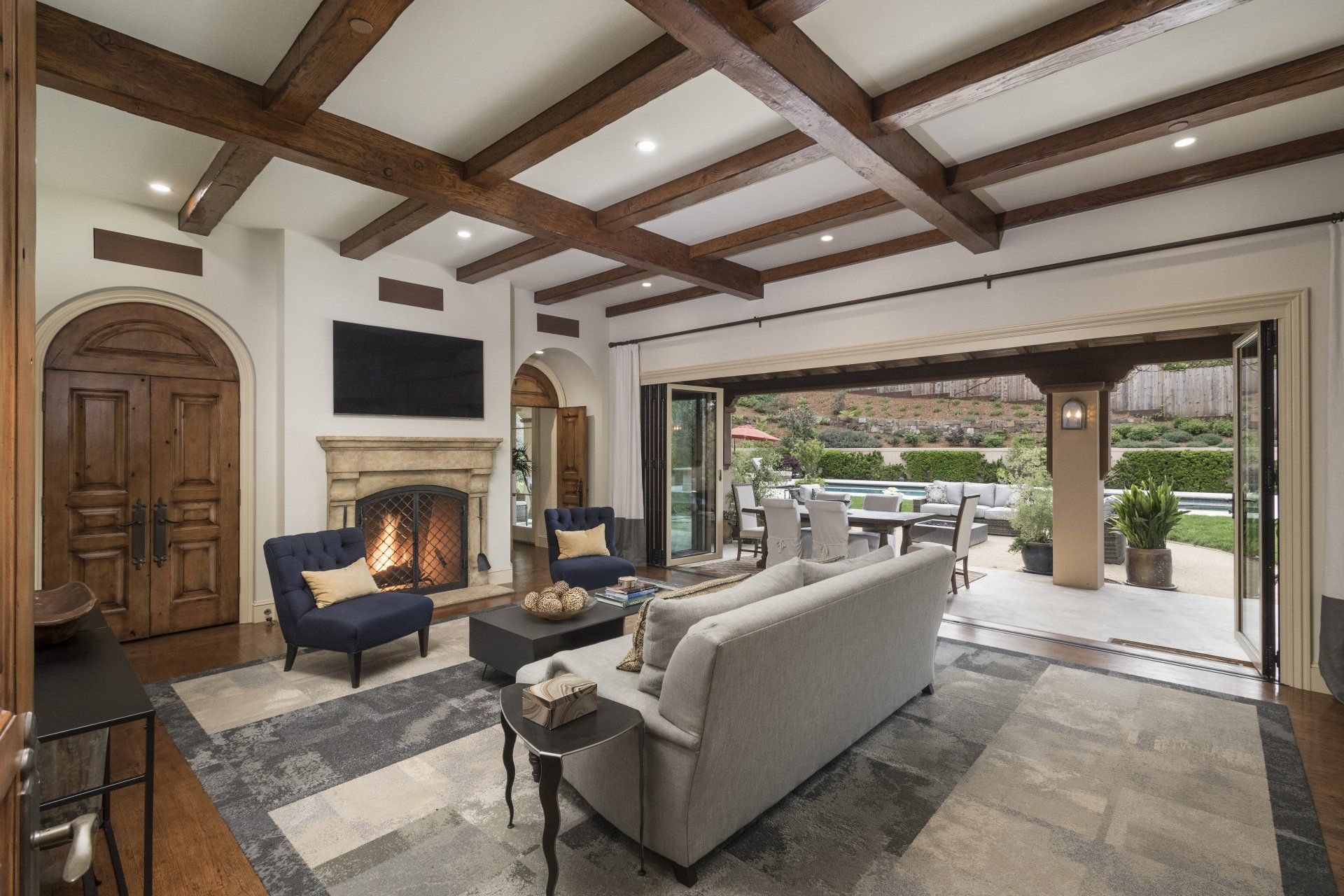 Living room with stone floor, fireplace, and open doors to patio. Wooden beams and furniture in neutral tones.