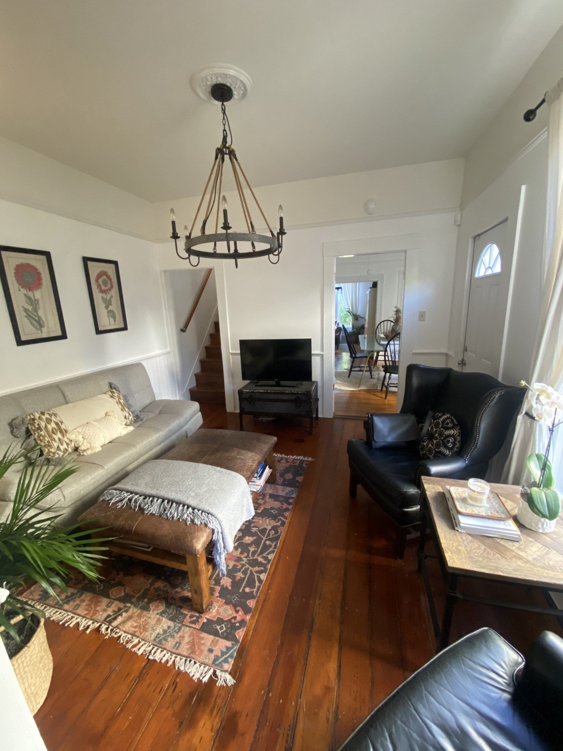 Cozy room with a sofa, ottoman, armchair, TV, and chandelier. Hardwood floors and white walls.