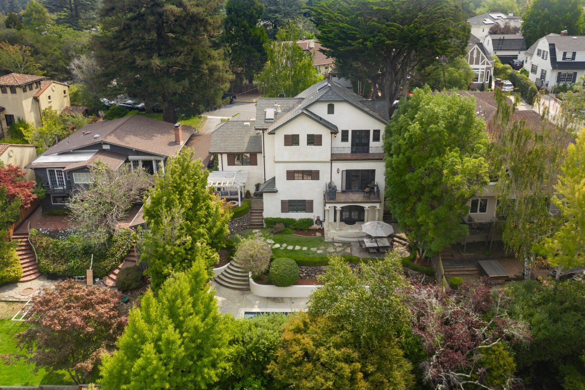 An aerial view of a large house surrounded by trees and other houses in a residential neighborhood.