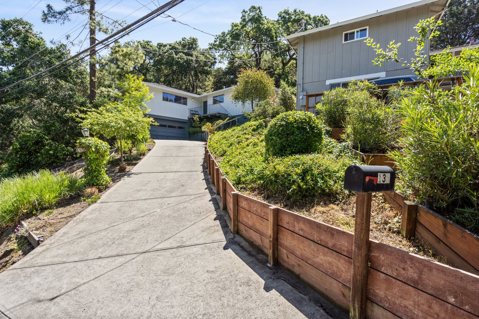 A driveway leading uphill towards the house; a wooden fence and green landscaping line the sides. A mailbox stands in the foreground.
