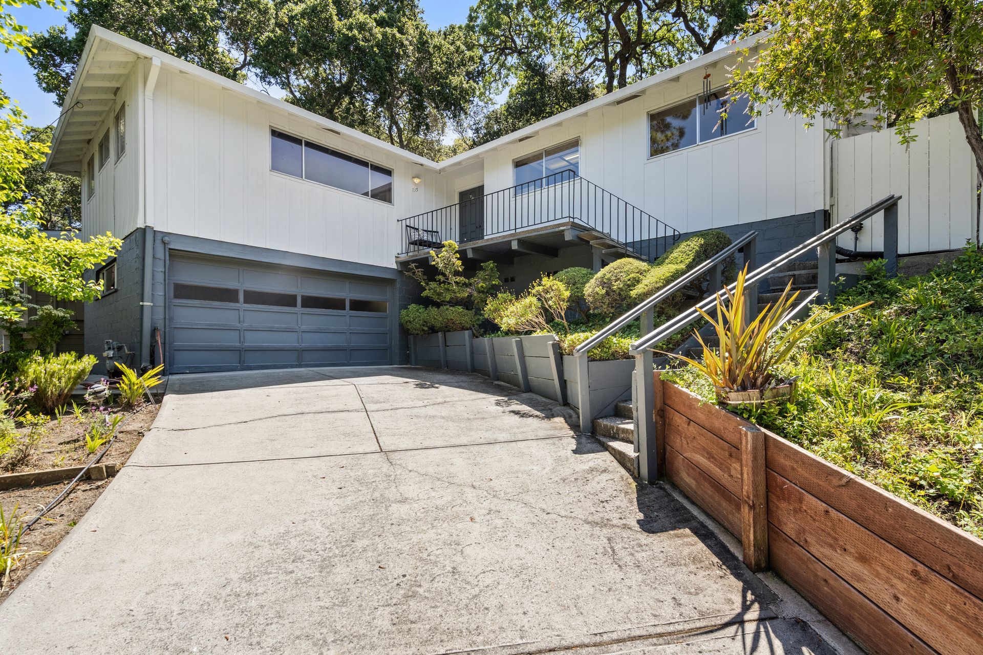 Two-story white house with gray garage door, concrete driveway, and wooden accents on a sloped lot.