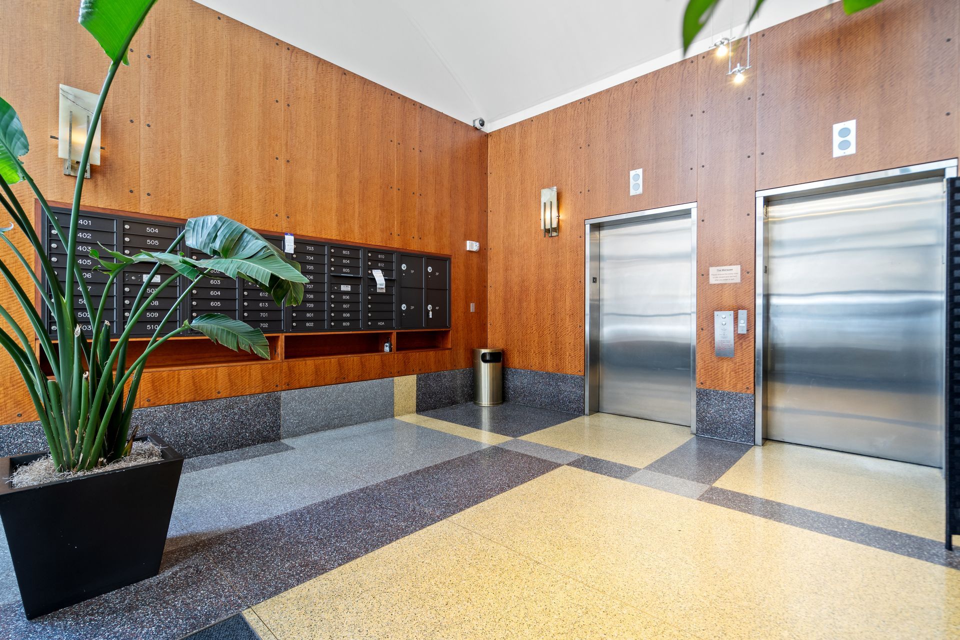 Lobby with two elevators, mailboxes, a large potted plant, and wood paneling. Floor has speckled tile.