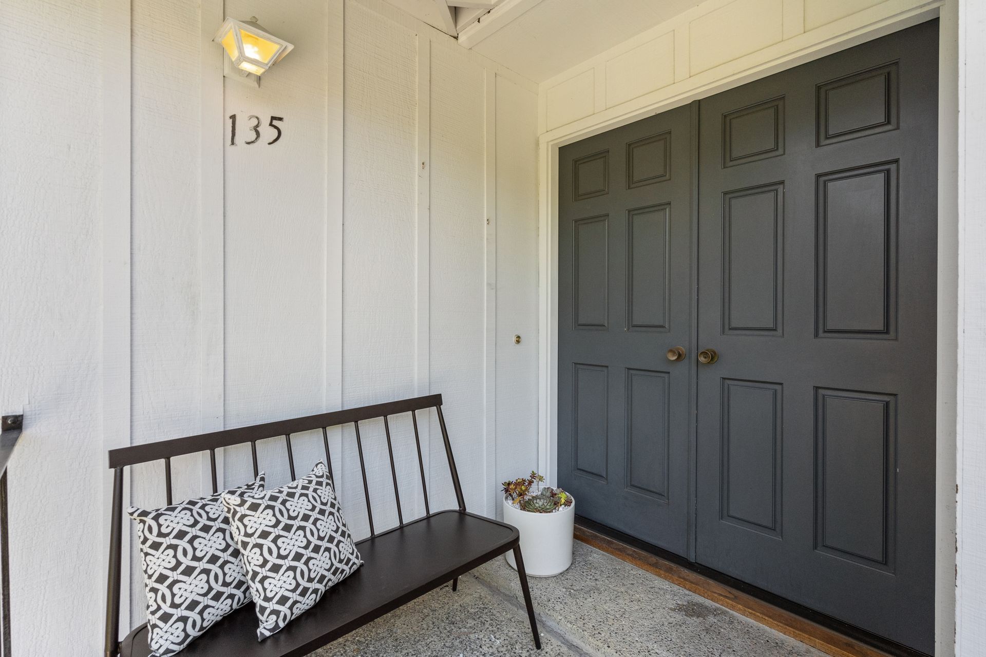 Exterior of a home entrance with dark gray double doors, a black bench with pillows, and the house number