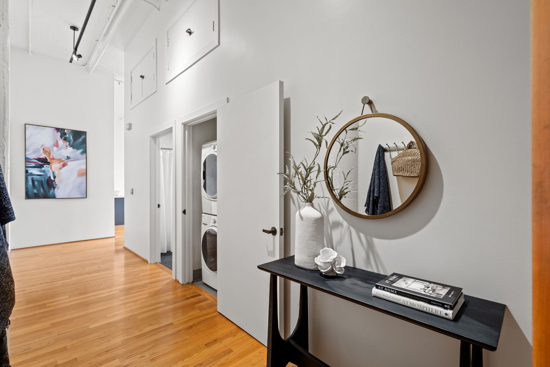 Hallway with hardwood floors, off-white walls, mirror, and decorative table.  Artwork and washing machine visible.