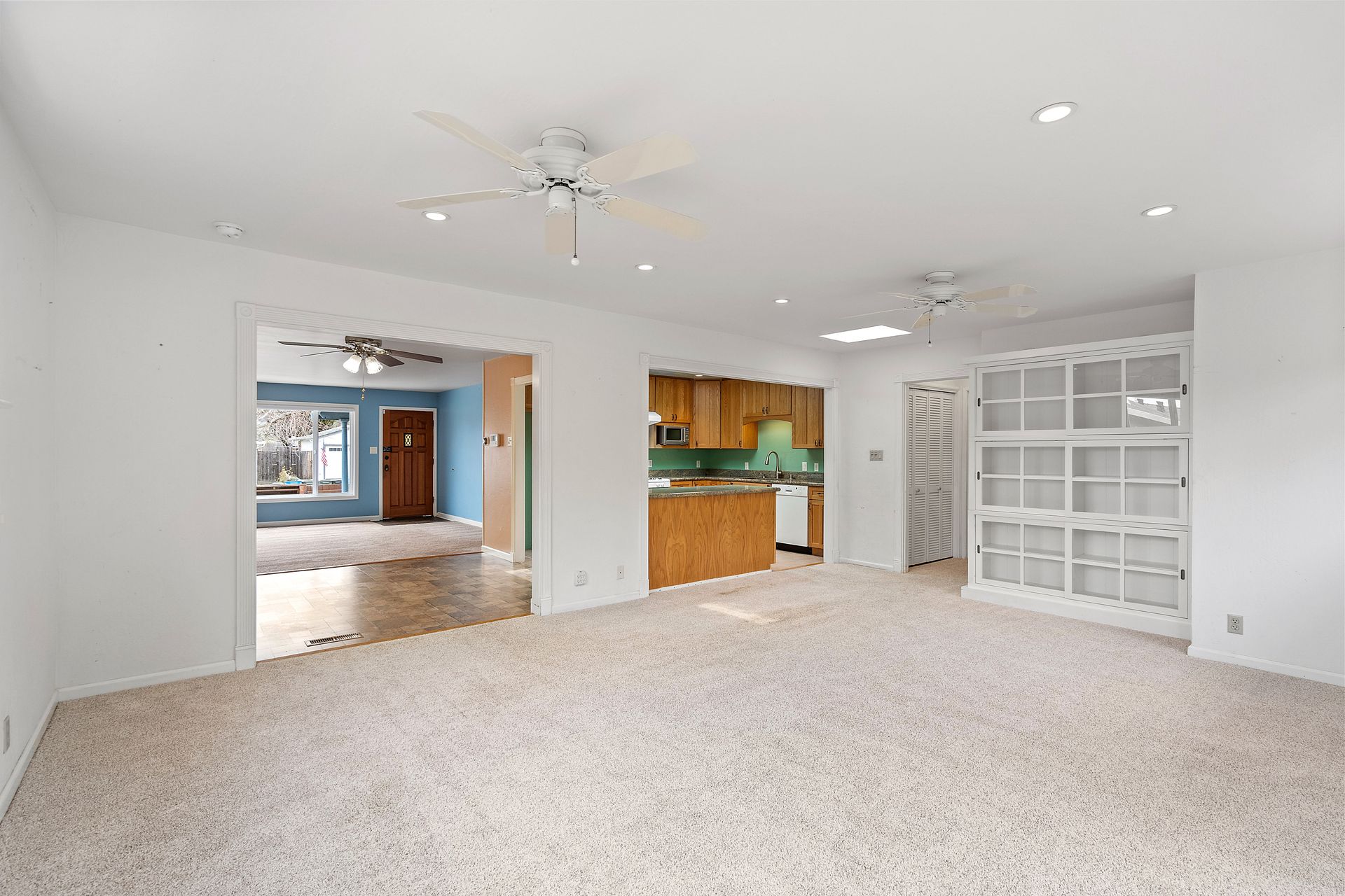 An empty room with a ceiling fan and a kitchen in the background.