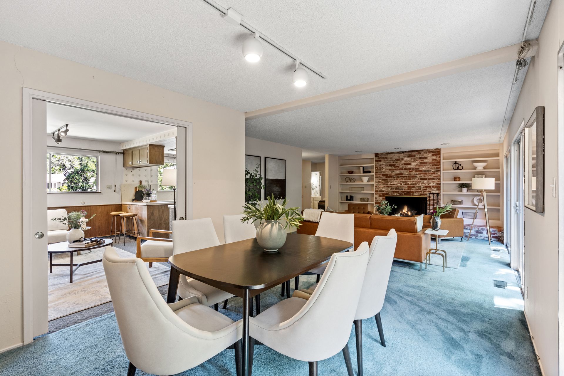 Dining room with a dark wood table, white chairs, and a view into the kitchen and living room with a brick fireplace.