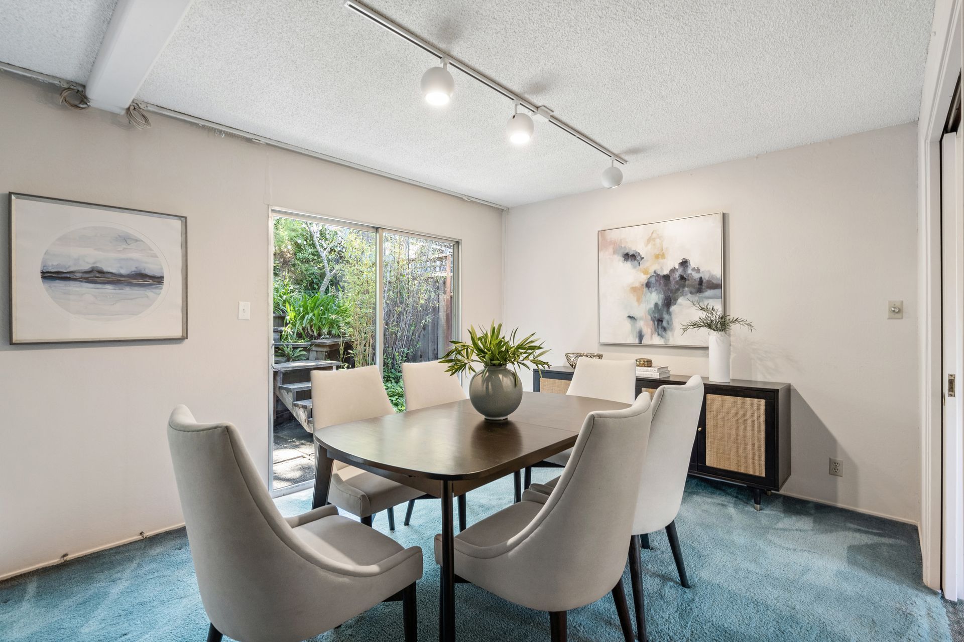 Dining room with a wooden table, six white chairs, a modern abstract painting, and a sliding glass door leading to greenery.
