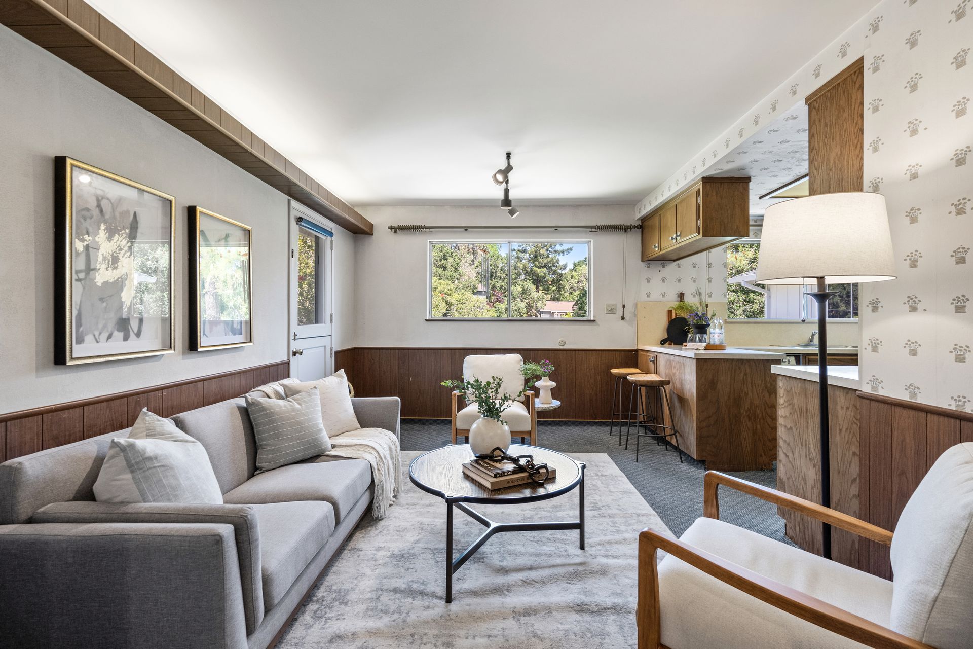 A room with a gray sofa, framed art, and light wood paneling. Kitchen visible in the background with matching wood cabinetry and a window.