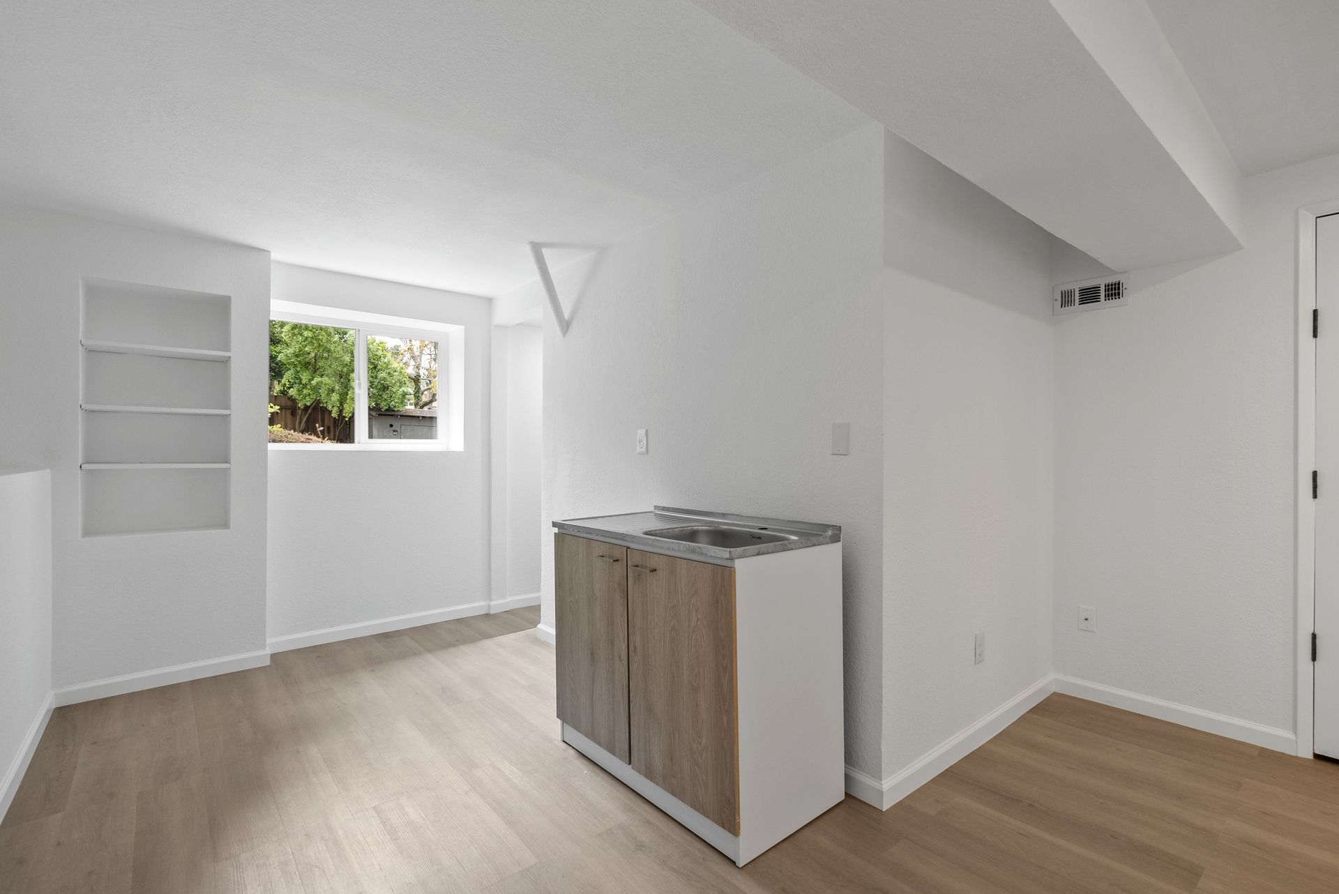 An empty room with hardwood floors , white walls , a sink and a window.