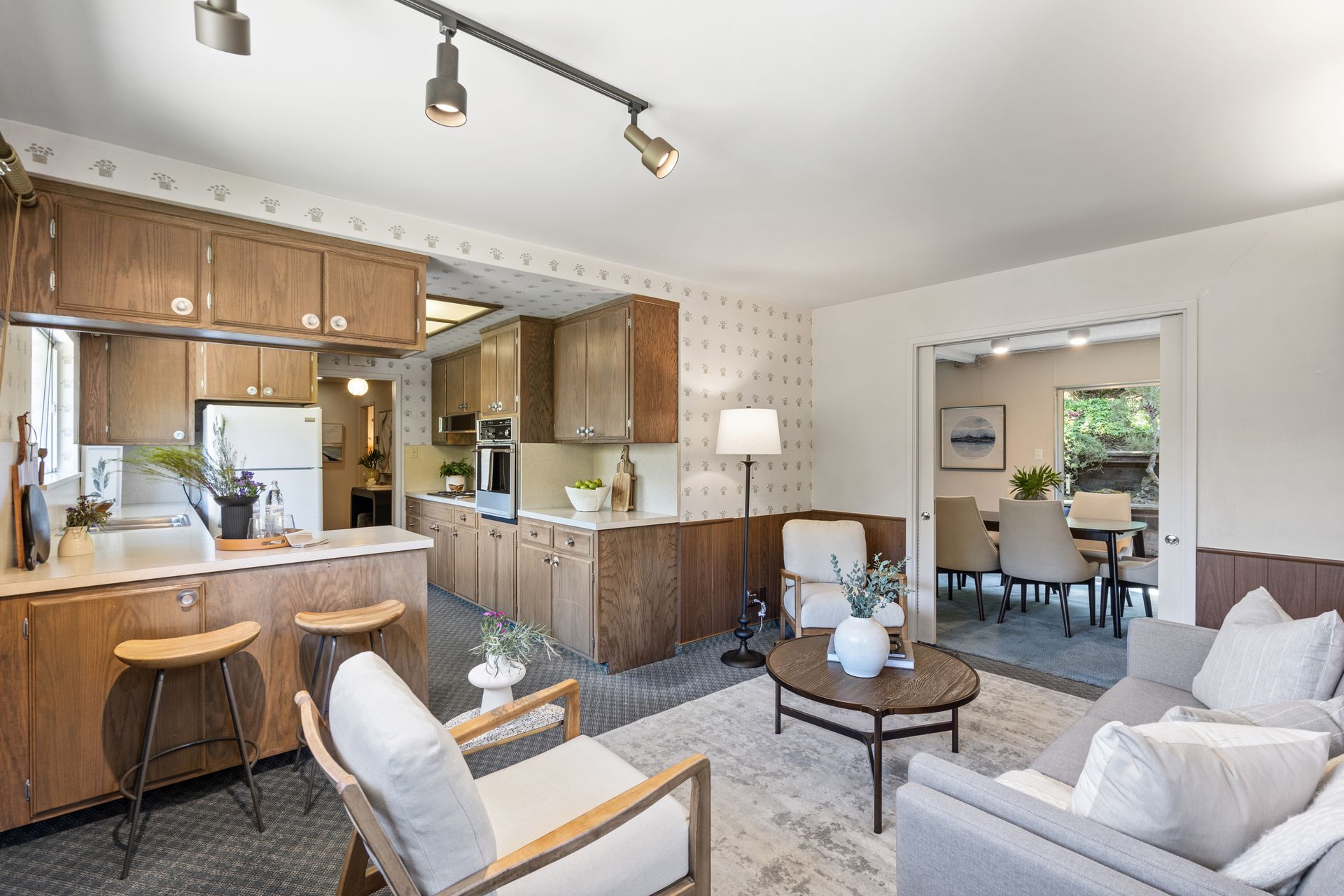 Open-concept living space with kitchen, dining area, and living room. Features wooden cabinets, a breakfast bar, and neutral-toned furniture.