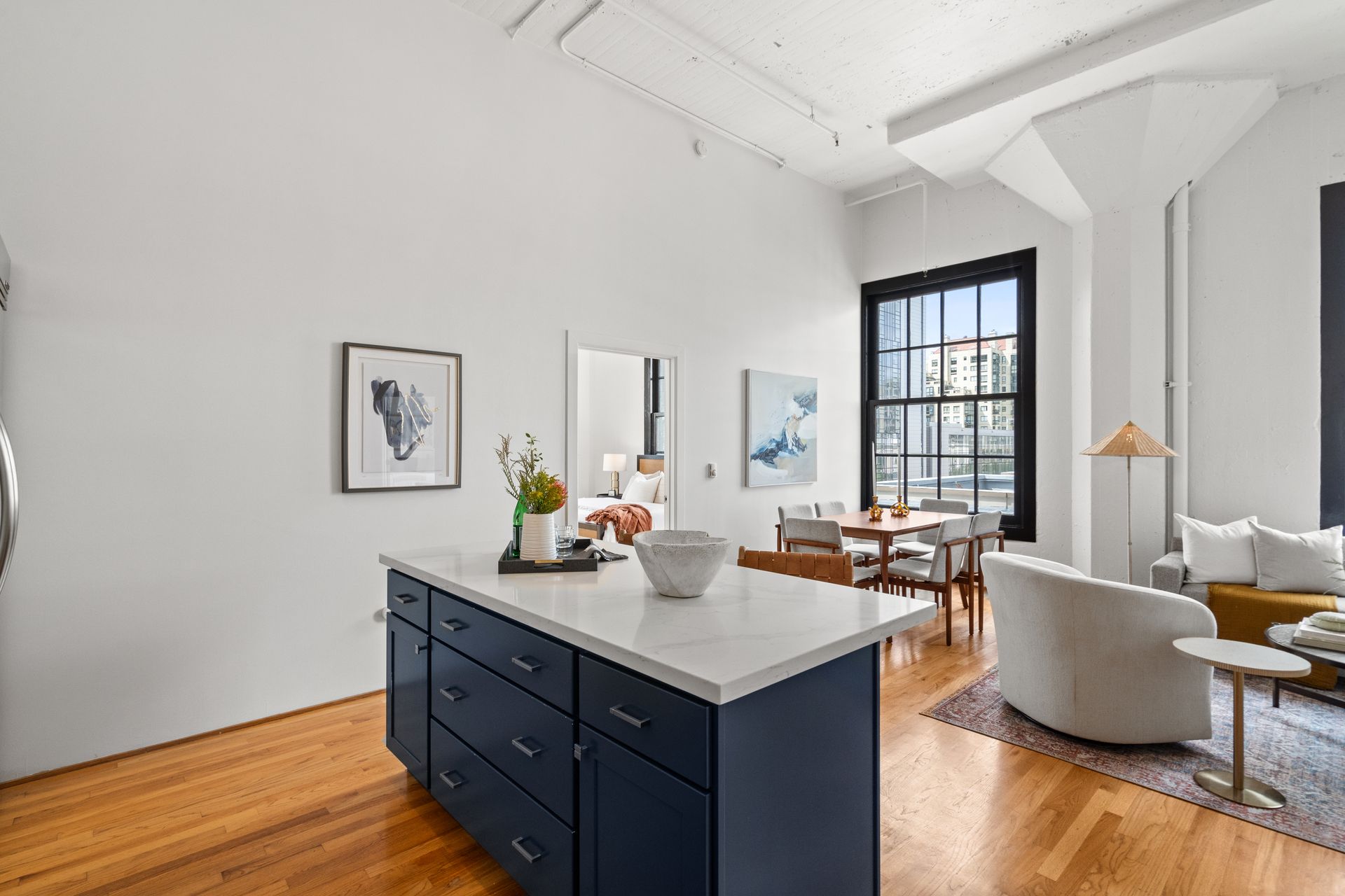 Open-concept kitchen with an island, hardwood floors, and dining area. Off-white walls, natural light, and modern furniture.