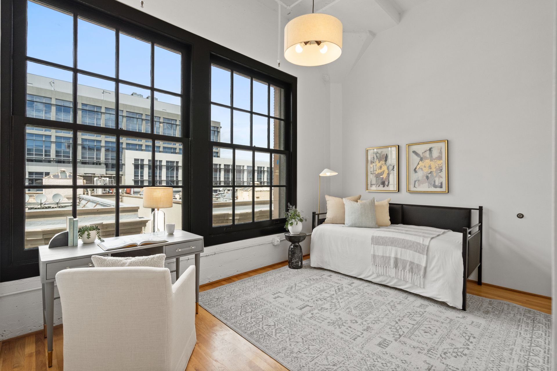A modern bedroom with a large window, desk, bed, and rug. The room is off-white with black window frames.