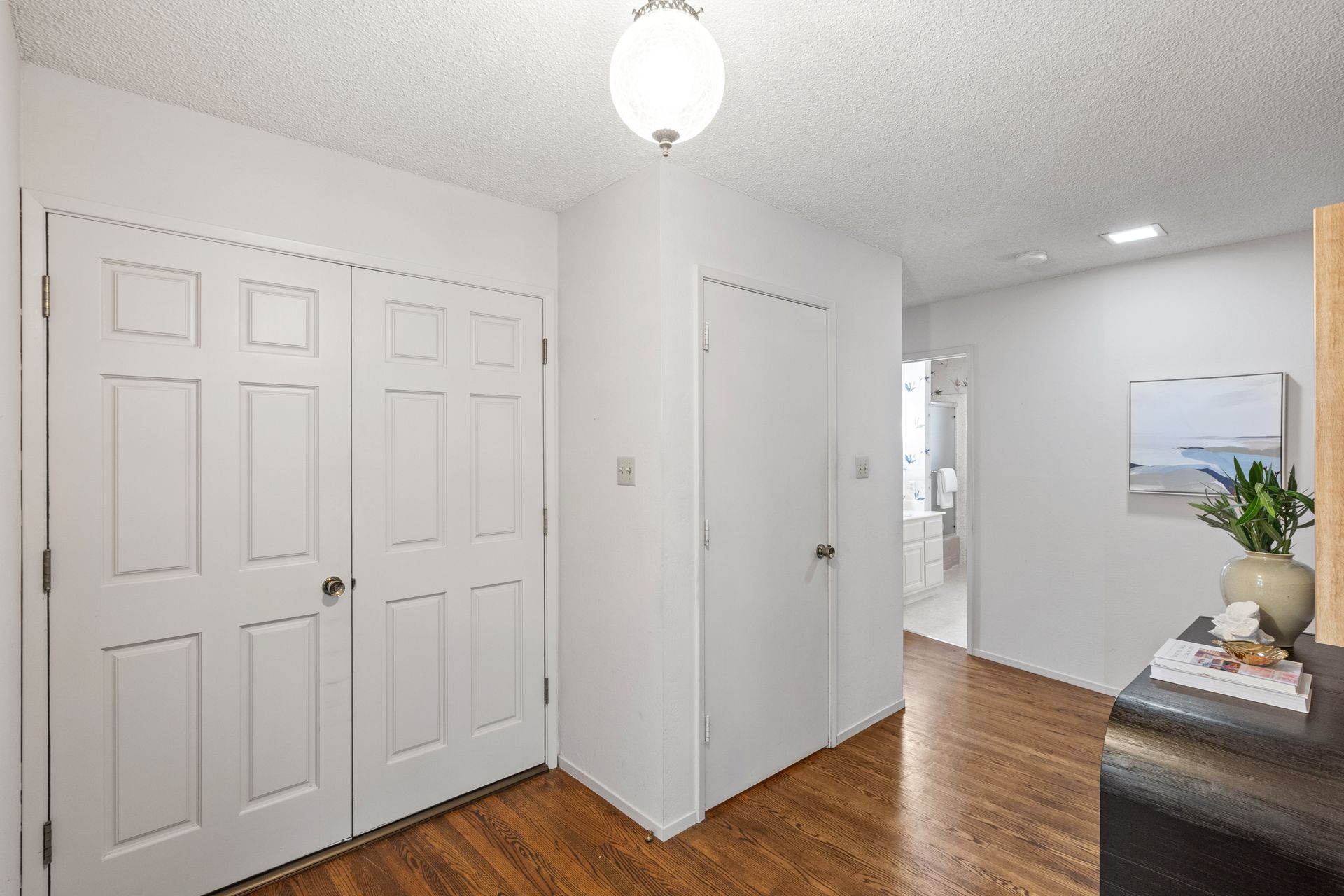 Hallway with white doors and walls, wood floors, and a small table with a painting and decor.