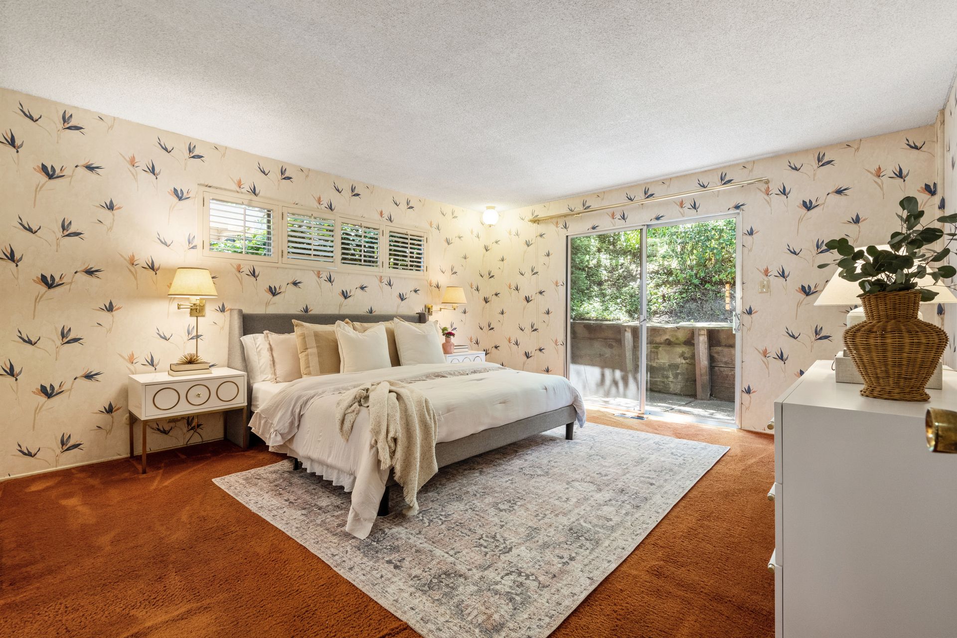 Bedroom with floral wallpaper, a bed with pillows and a blanket, and a sliding door to a balcony. Orange-brown carpet and furniture are also present.