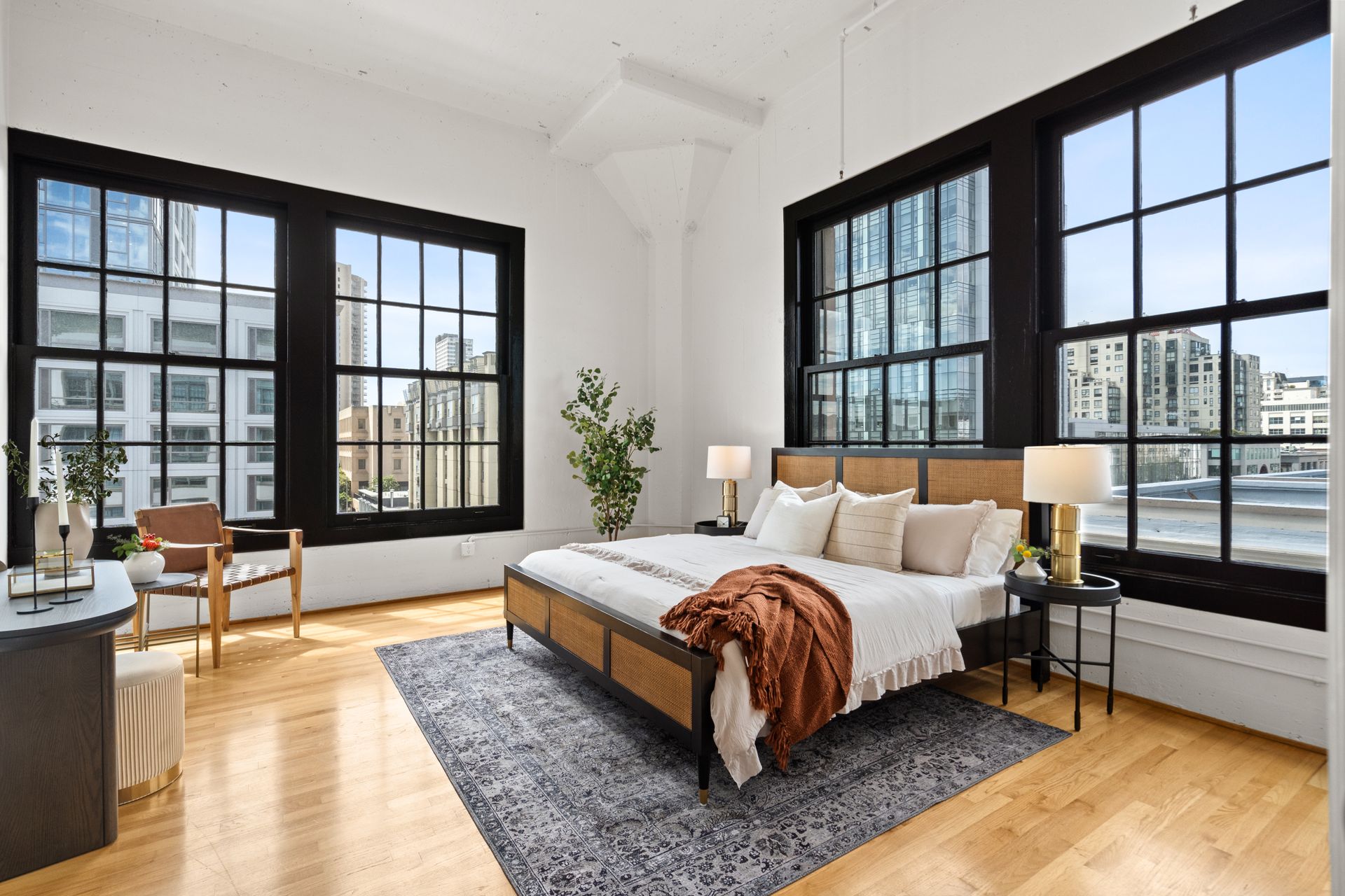 Spacious bedroom with large windows, light wood floors, bed, rug, and city views.
