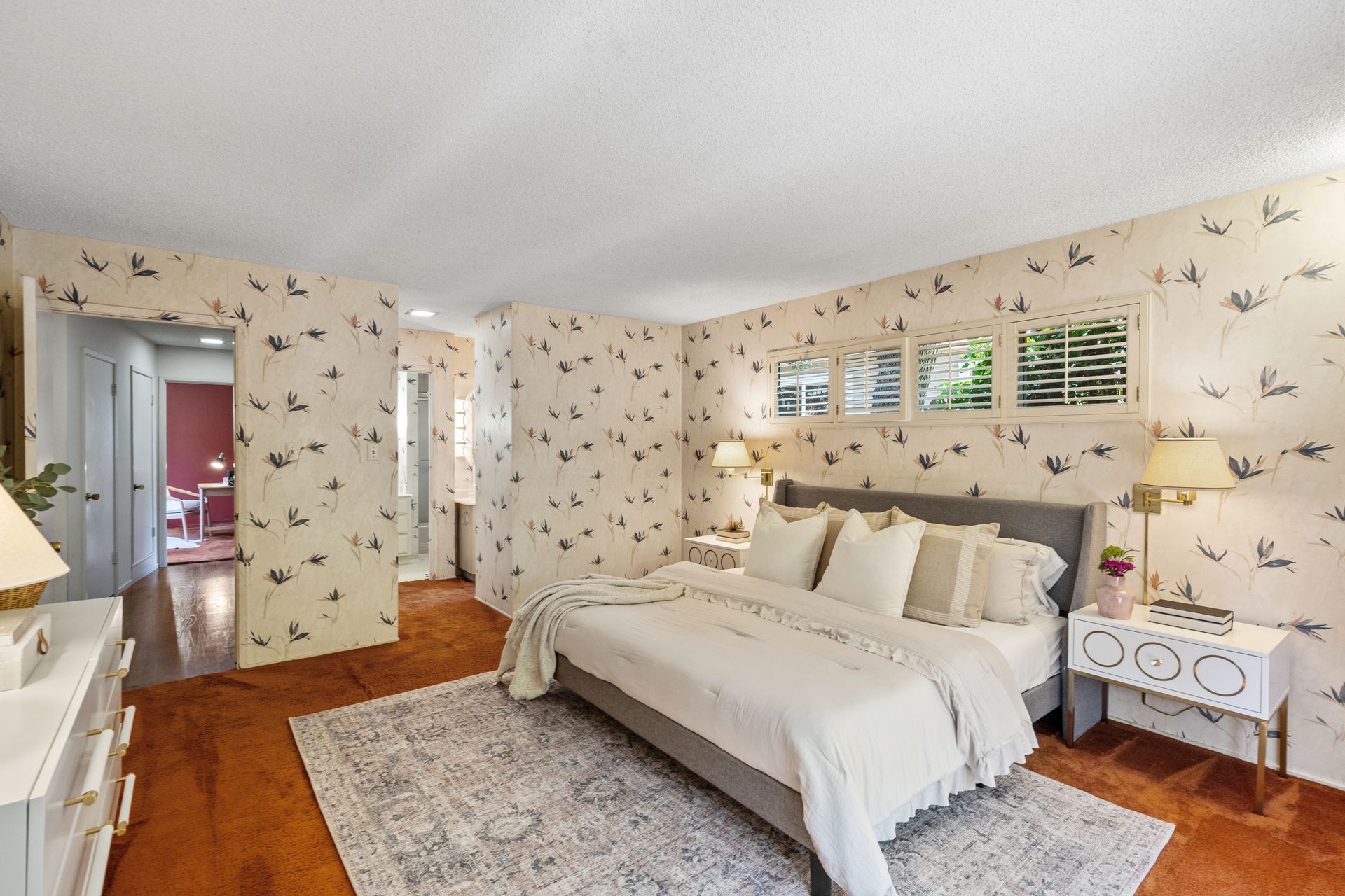 Bedroom with floral wallpaper, gray bed, white nightstand, and a patterned rug on an orange-brown carpet.