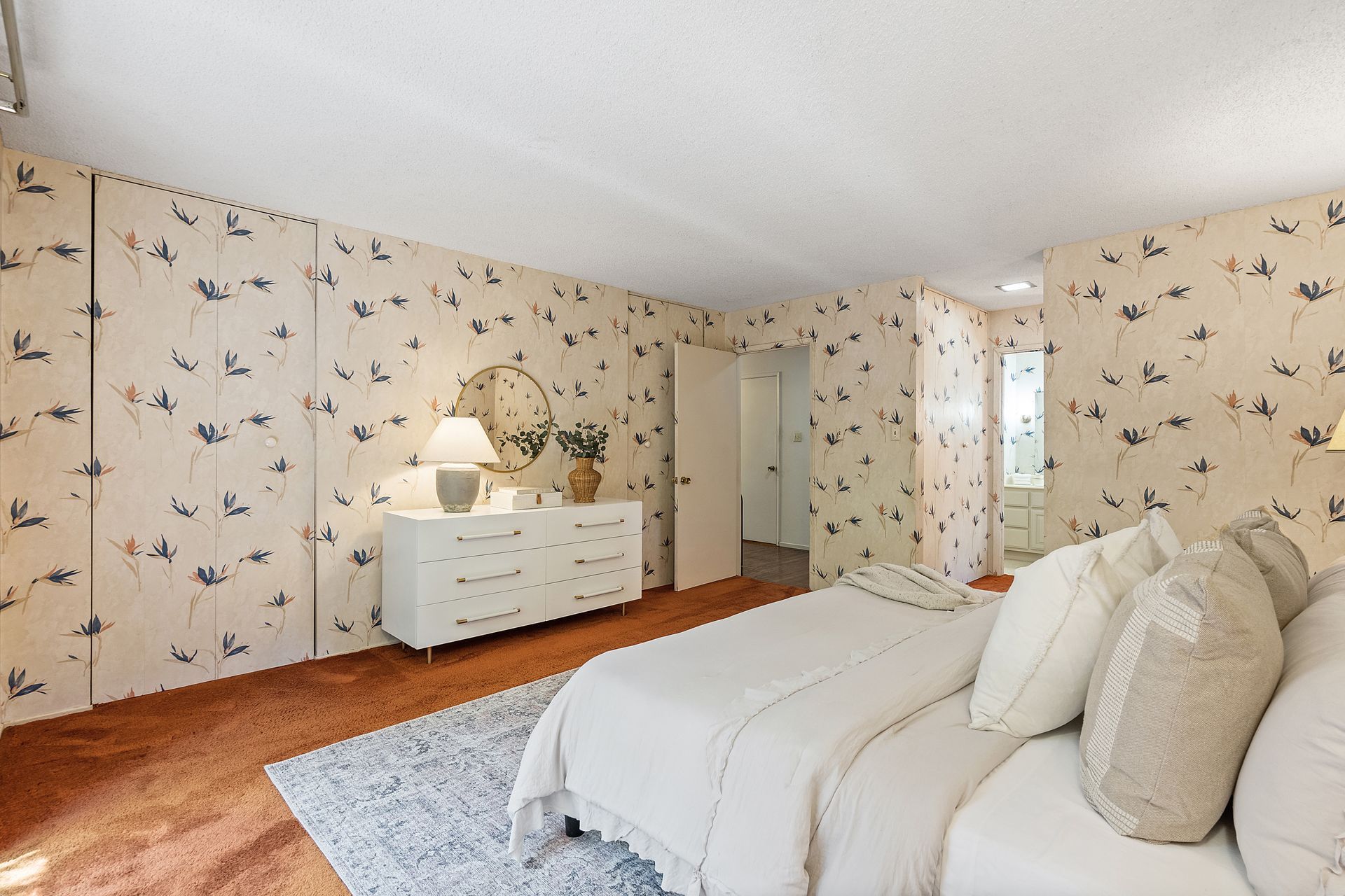 Bedroom with white furniture, floral wallpaper, and a bed with many pillows. The room has orange-brown carpeting.