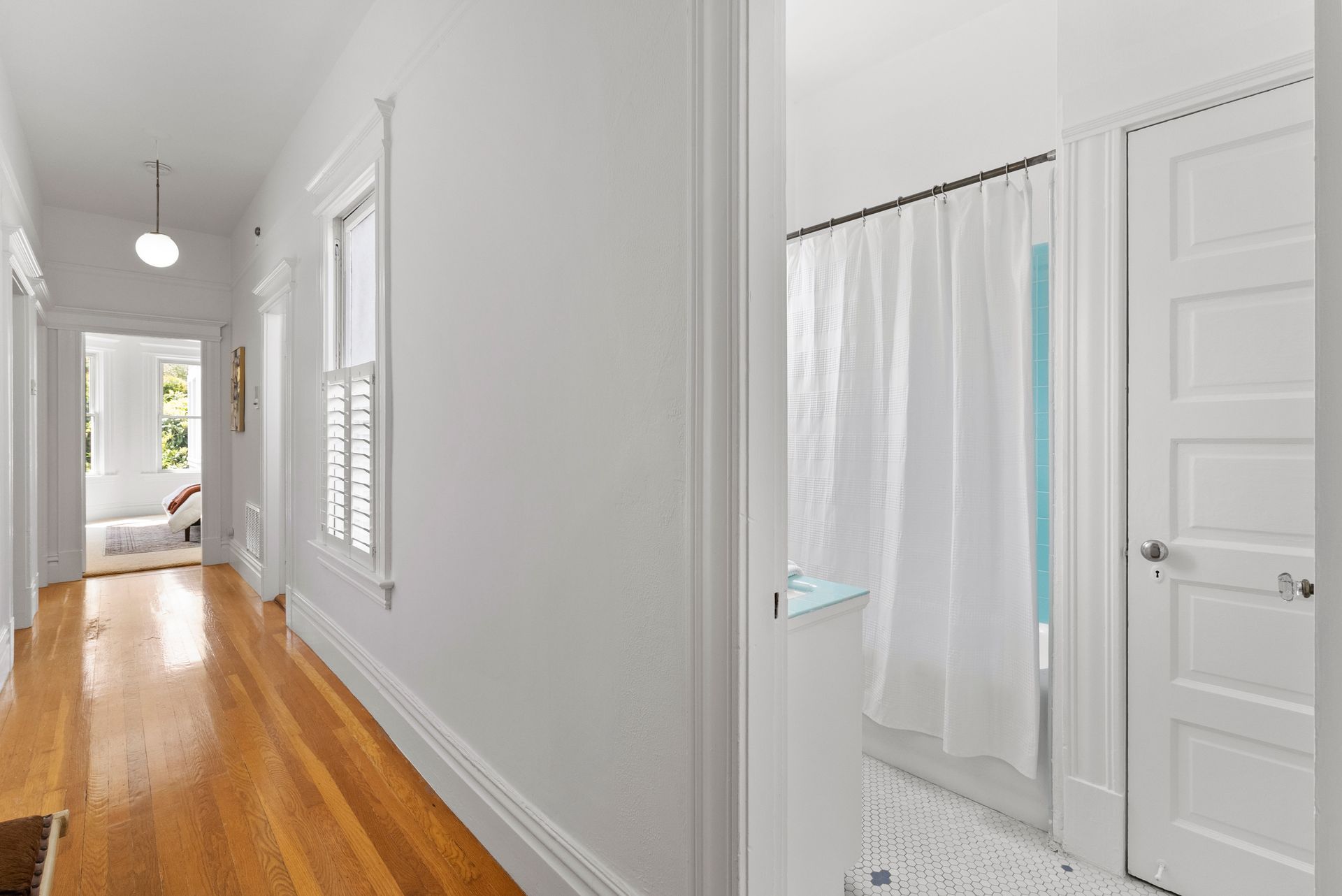 A long hallway with wooden floors and white walls leading to a bathroom.