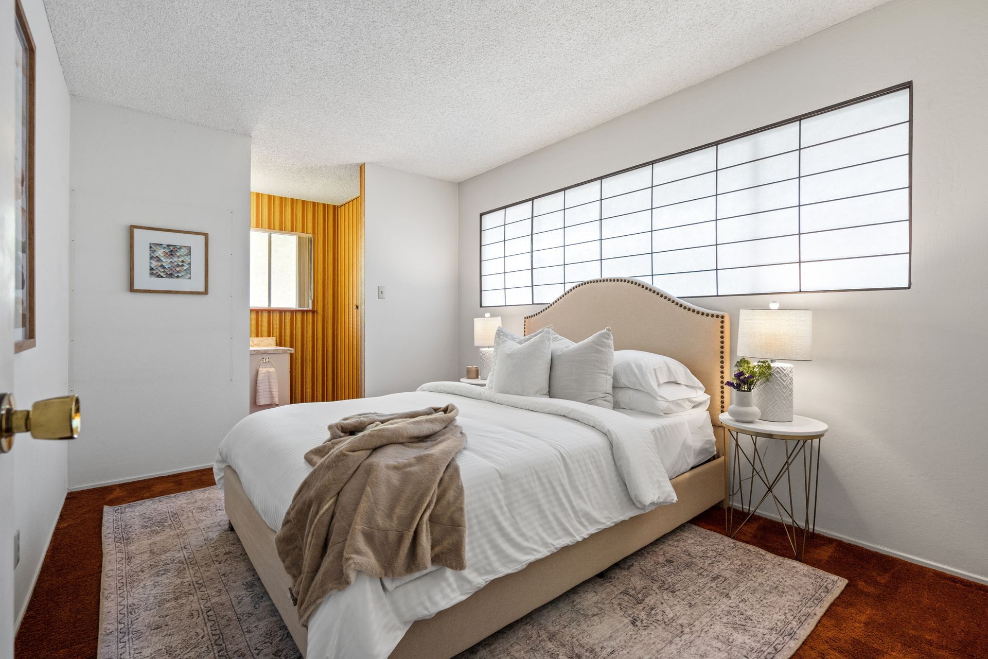 A bright bedroom with a bed, two side tables, and a large window. The room is white and has a tan bedspread and rug.