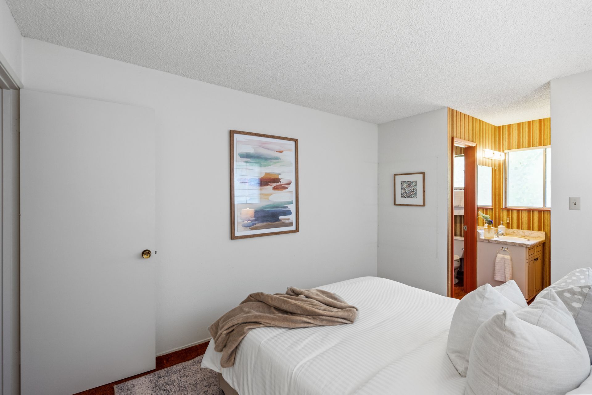 Bedroom with white walls, a bed, and a doorway leading to a bathroom with wood paneling. Artwork hangs on the wall.