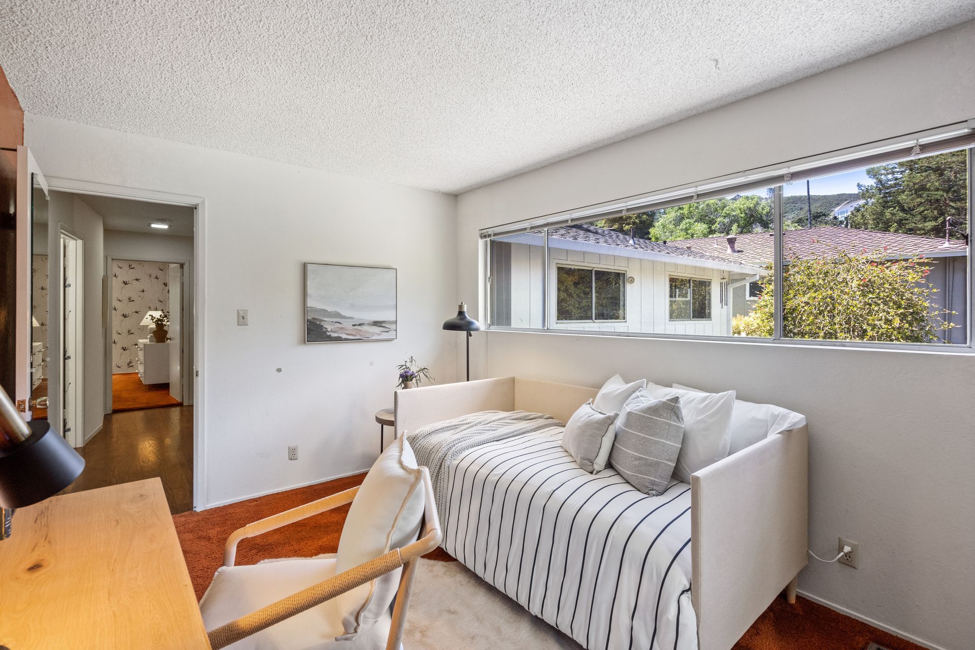 A room with a daybed, desk, and a large window overlooking greenery. The room has white walls and warm-toned accents.