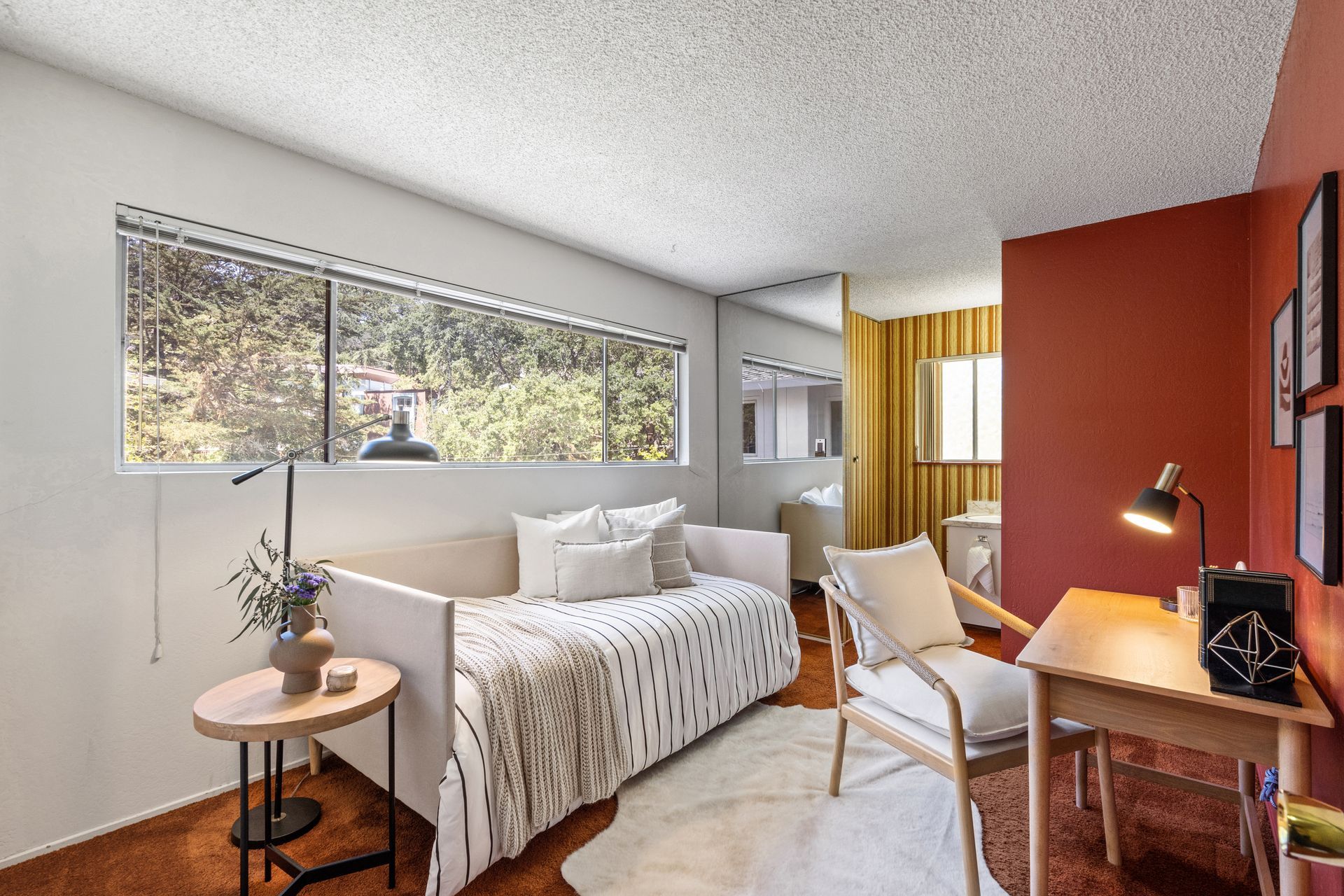 A room with daybed, desk, and chair. Red accent wall, window, and hardwood floors.