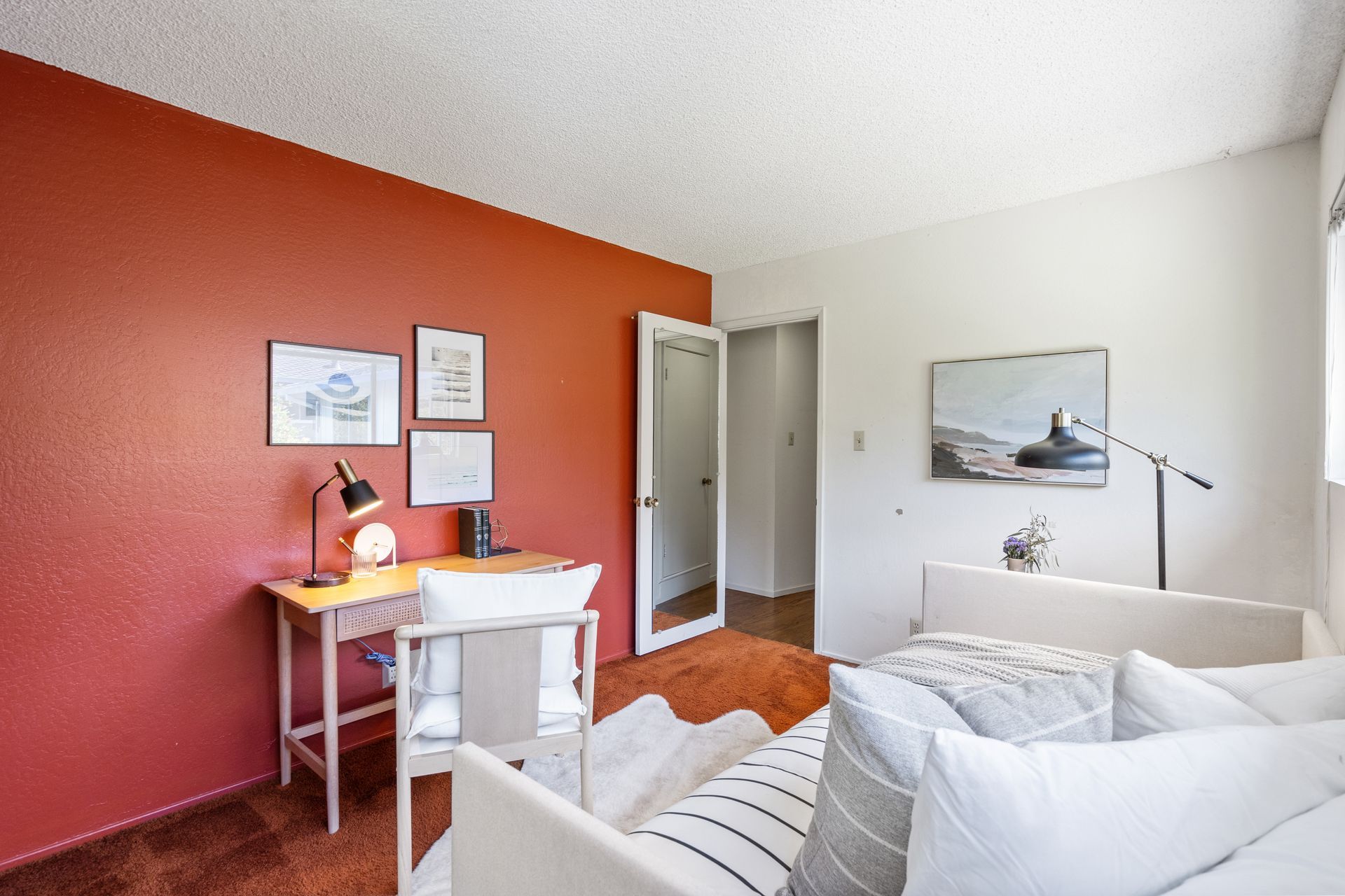 A room with an office area, a couch, and a red accent wall. A desk with a laptop and framed artwork is in front of the red wall.