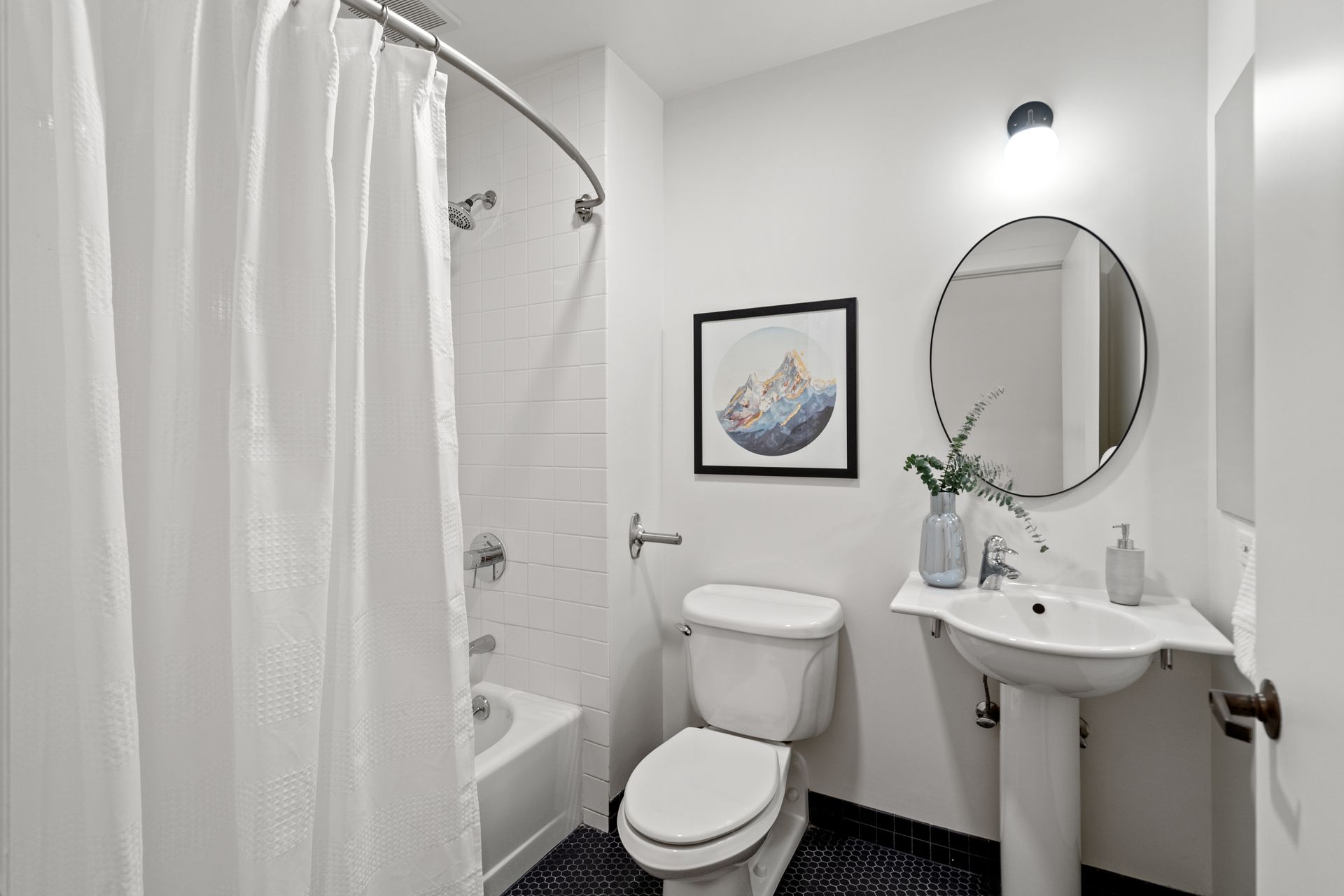 White bathroom with shower, toilet, pedestal sink, round mirror, and framed art.